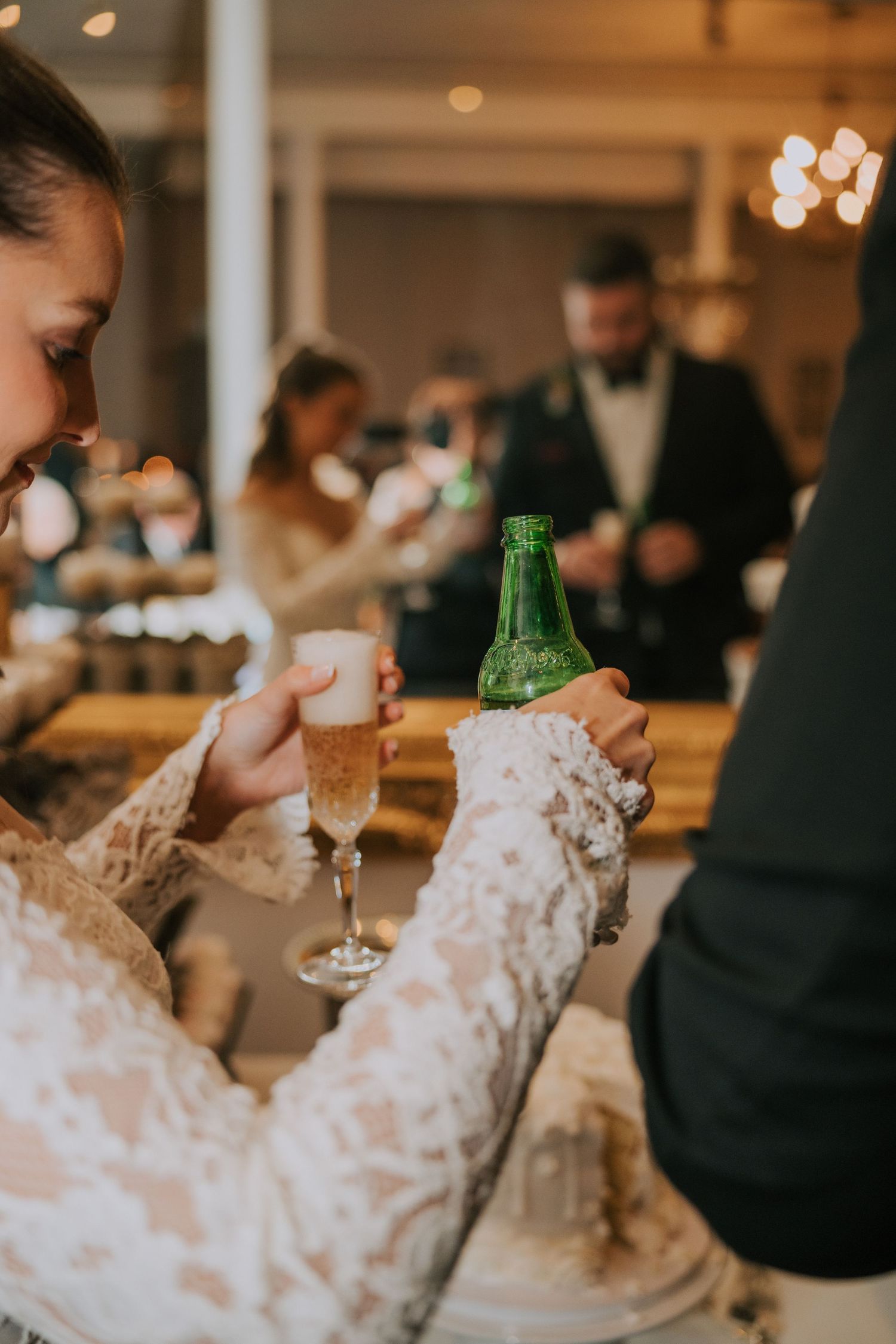 Elegant lace sleeve raises a beer bottle at a wedding reception with bokeh lights and guests in the background.