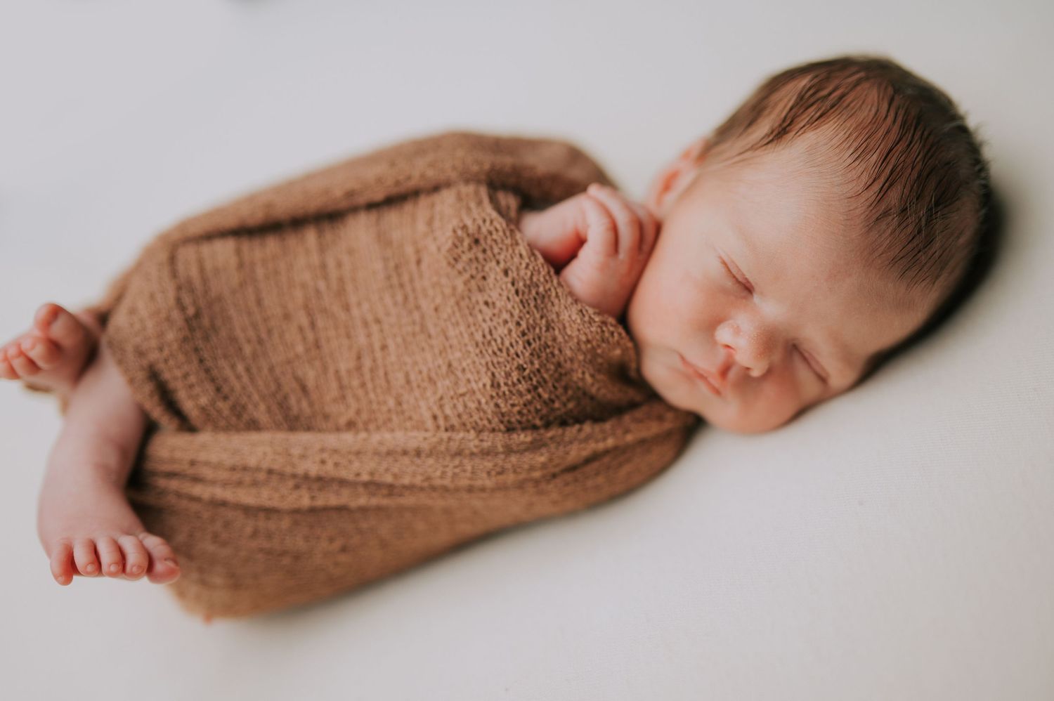 Sleeping newborn wrapped in brown knit blanket on white background.