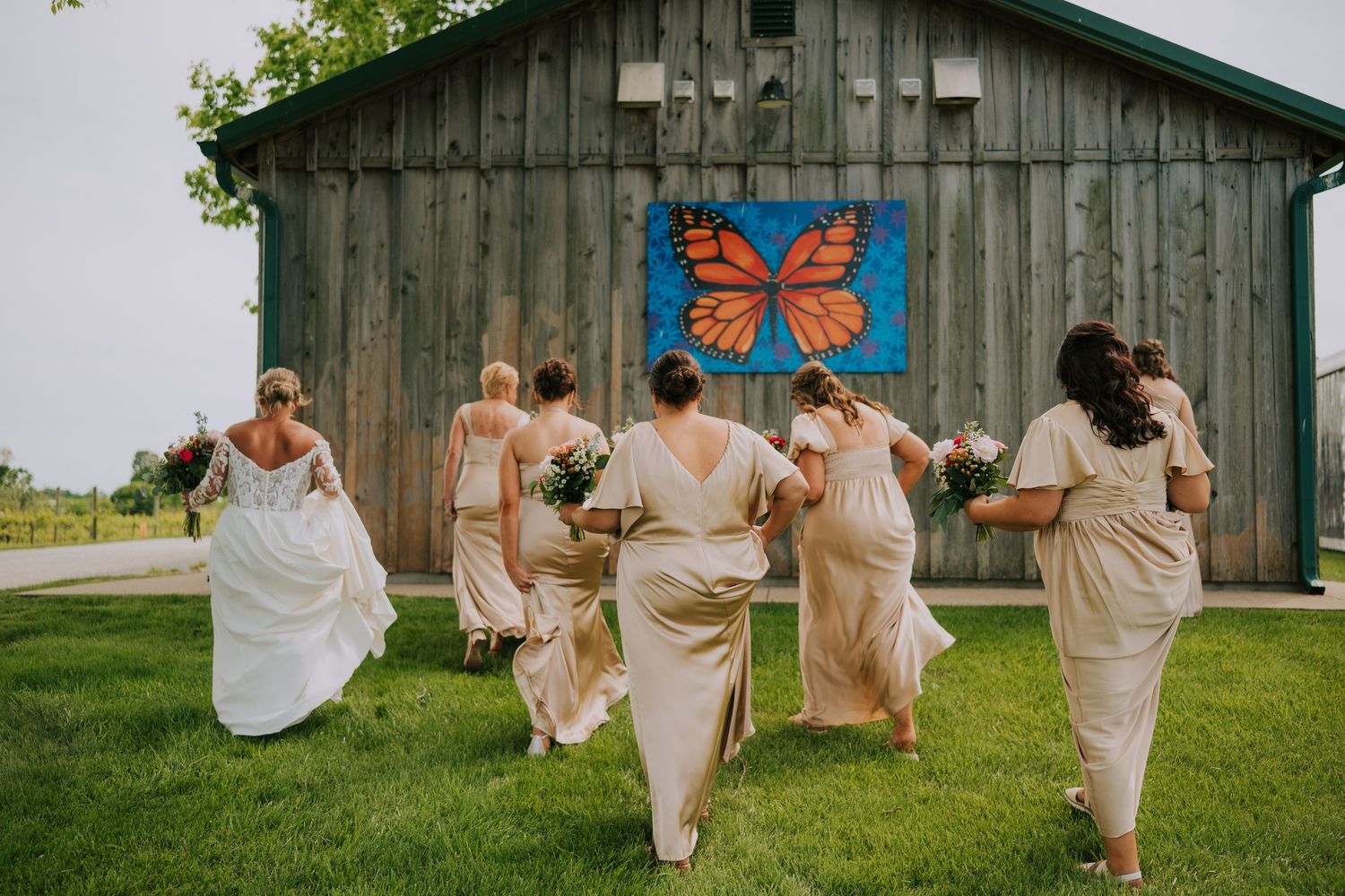 Bridesmaids in champagne dresses running playfully in front of rustic barn with butterfly artwork.