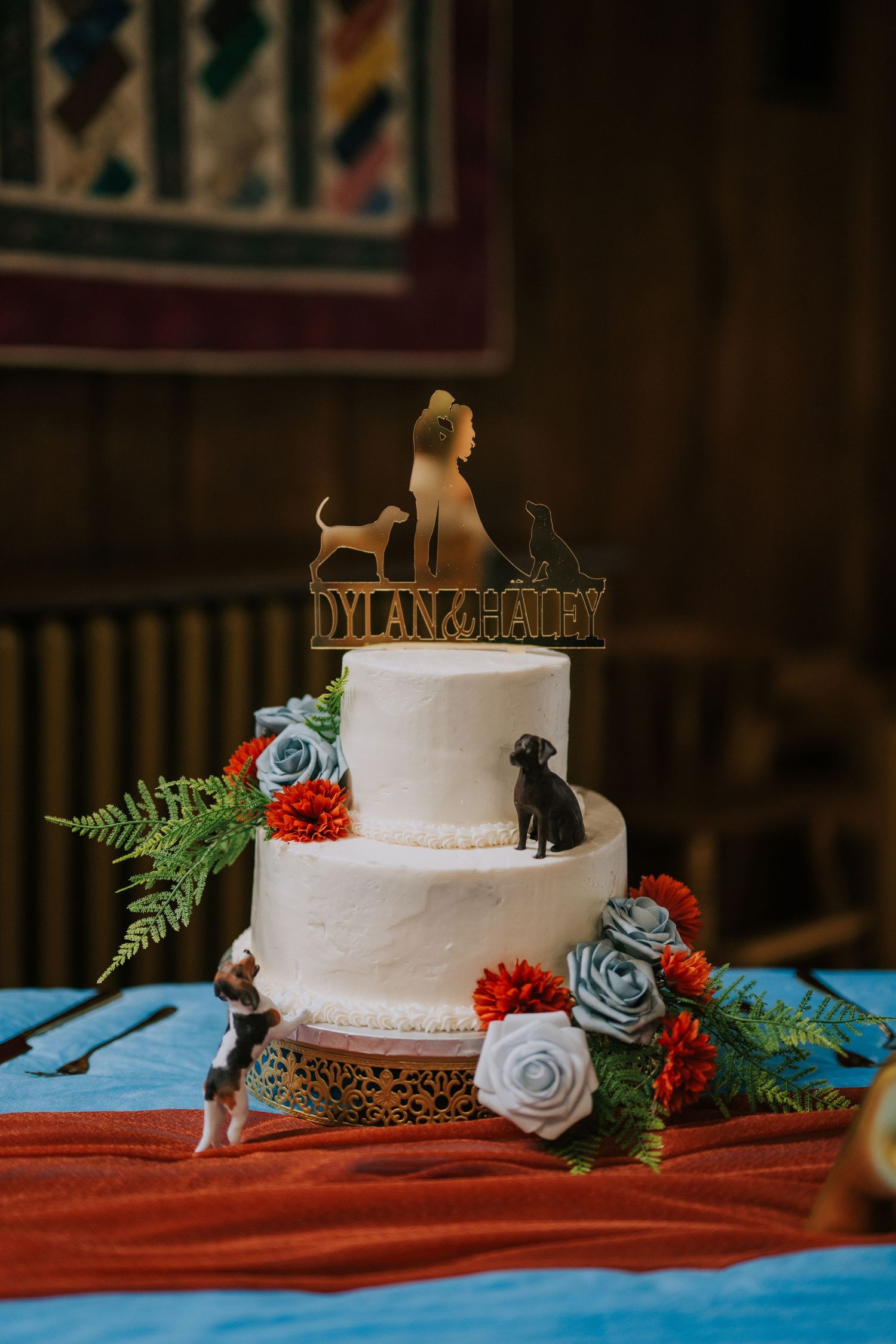 White wedding cake decorated with blue roses, ferns, and custom dog figurine cake toppers.