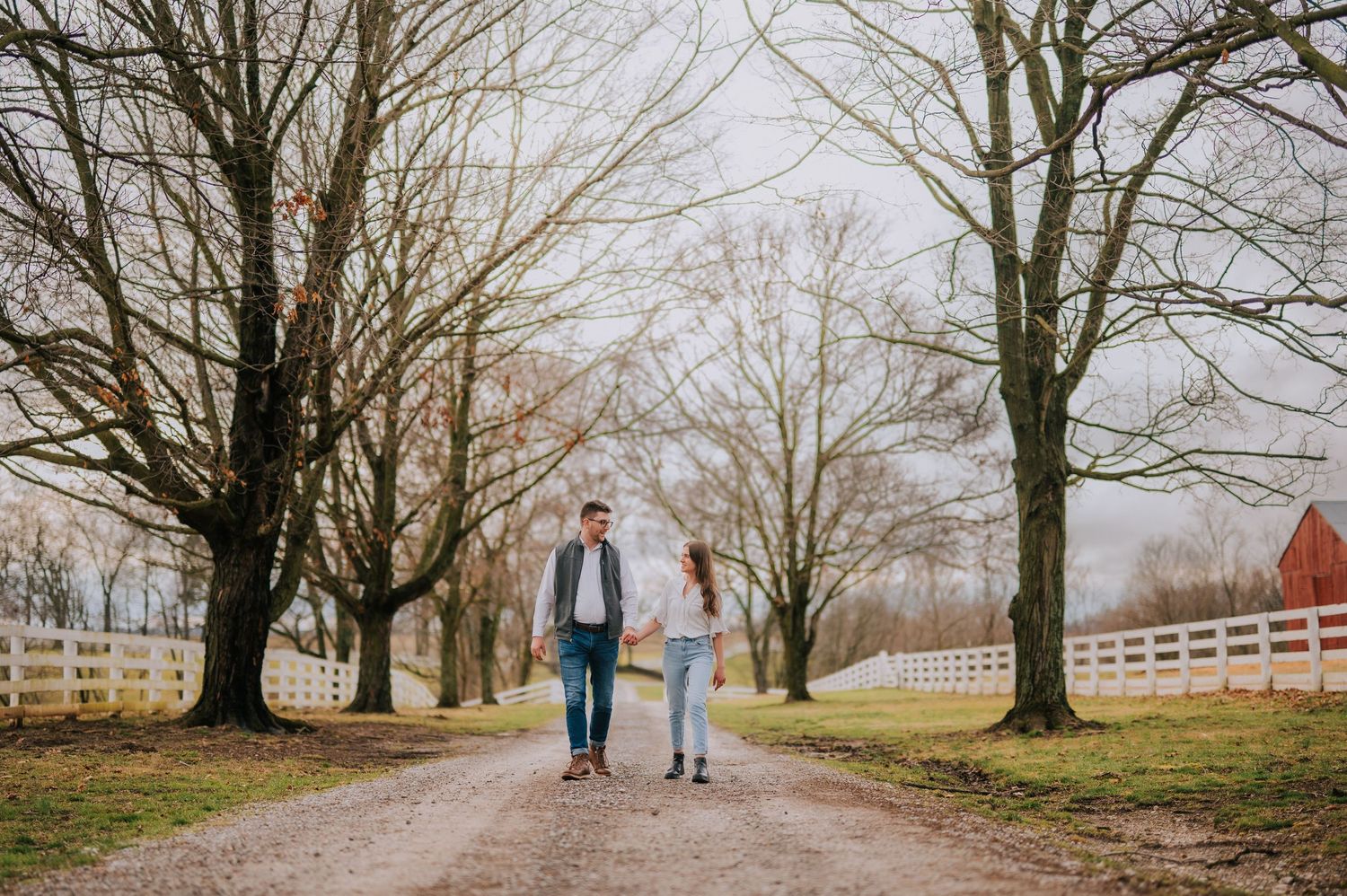 A couple strolls down a tree-lined path with a red barn visible in the background.