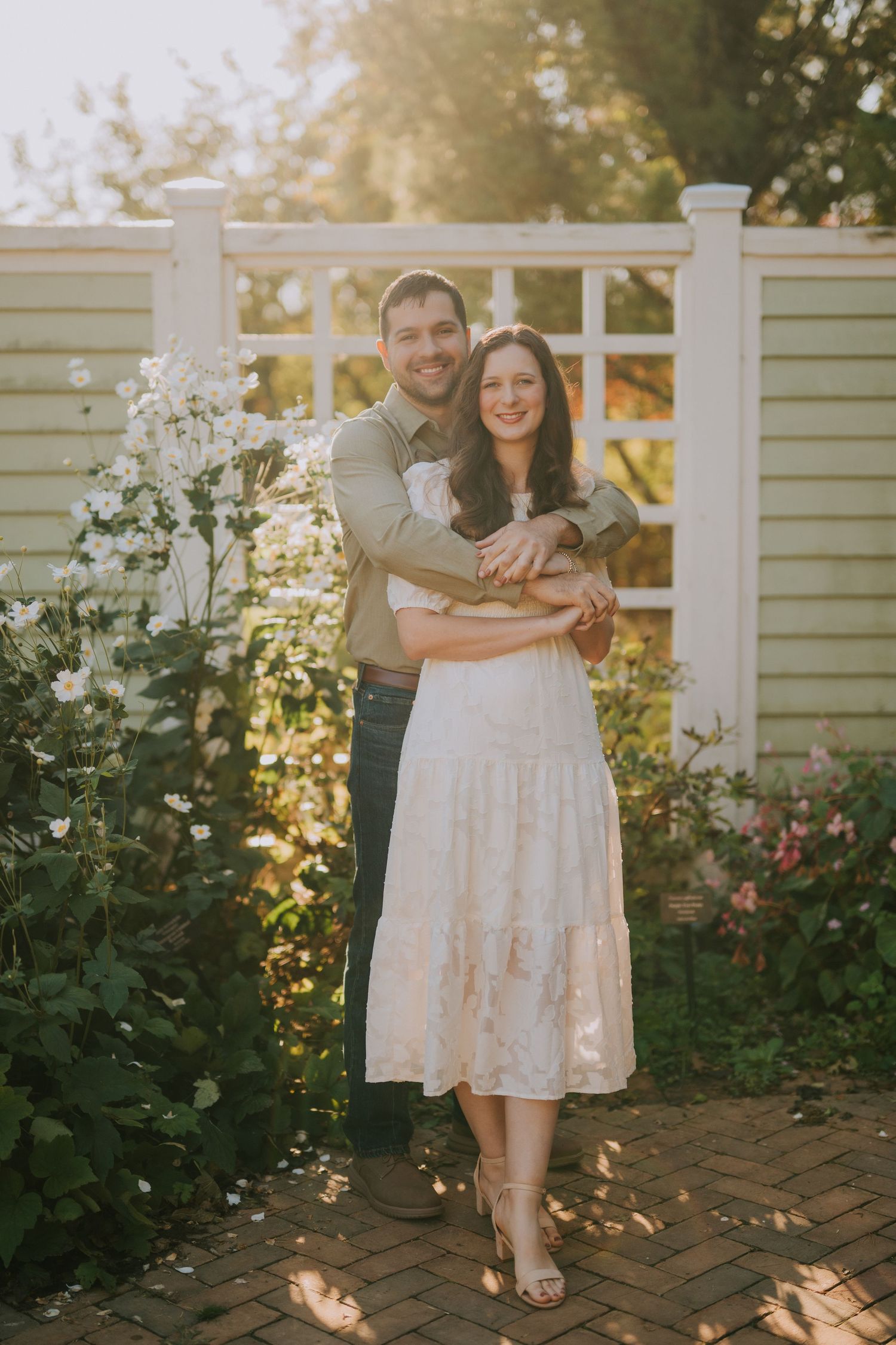 A couple poses on a garden deck with white railings and blooming flowers during sunset.