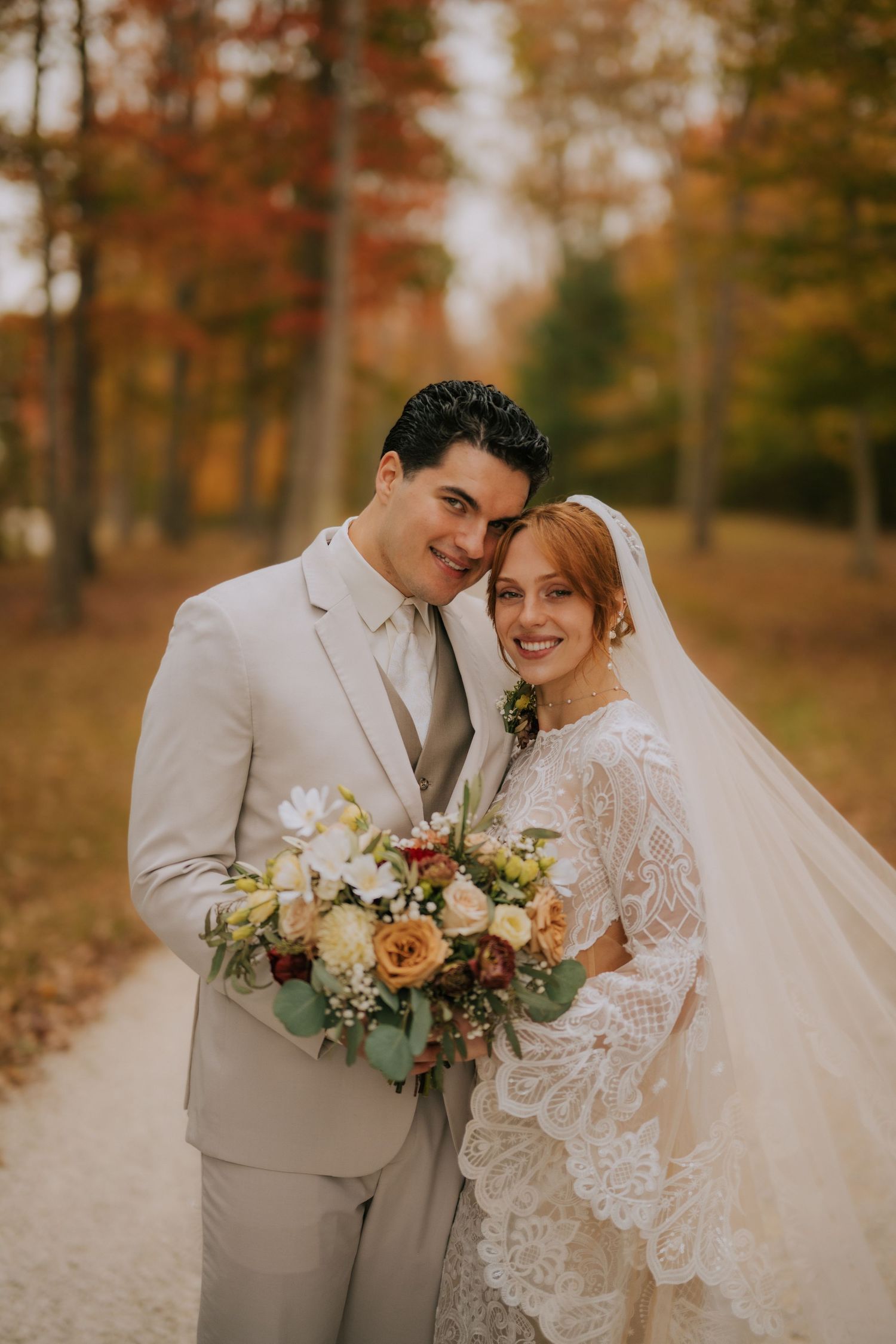 A smiling bride and groom pose together on an autumn path, she holds a bouquet in a lace gown with veil.