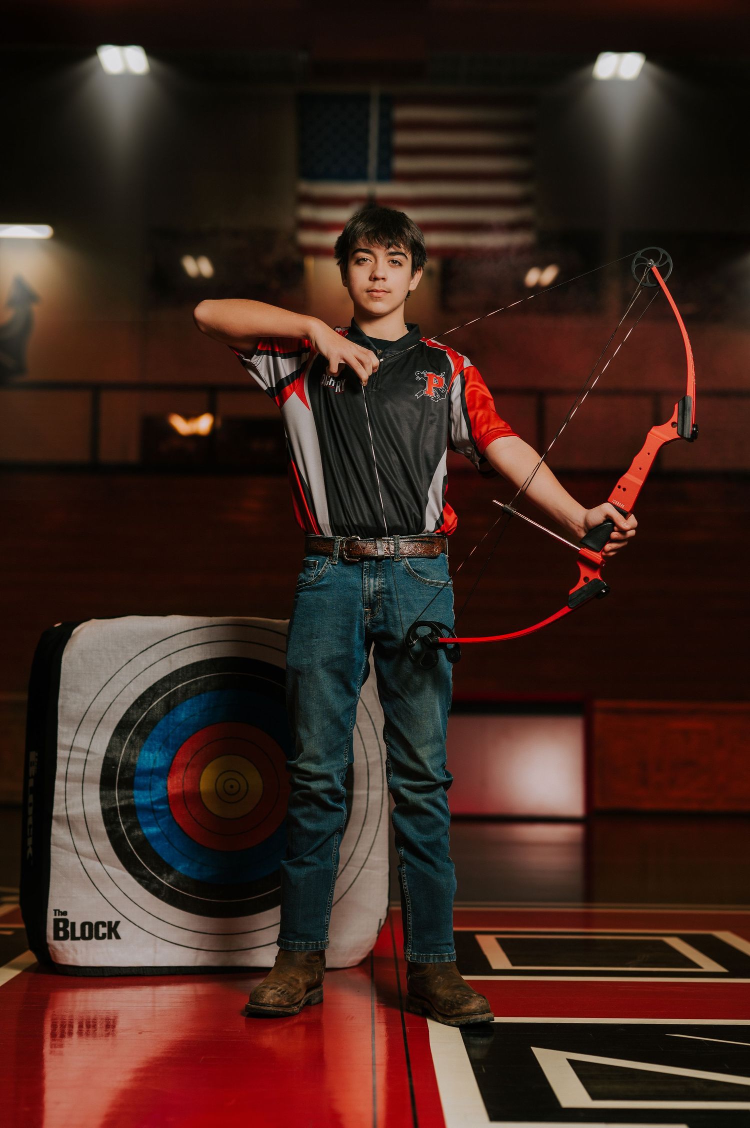 Young archer poses with bow and arrow in front of archery target during indoor practice session.