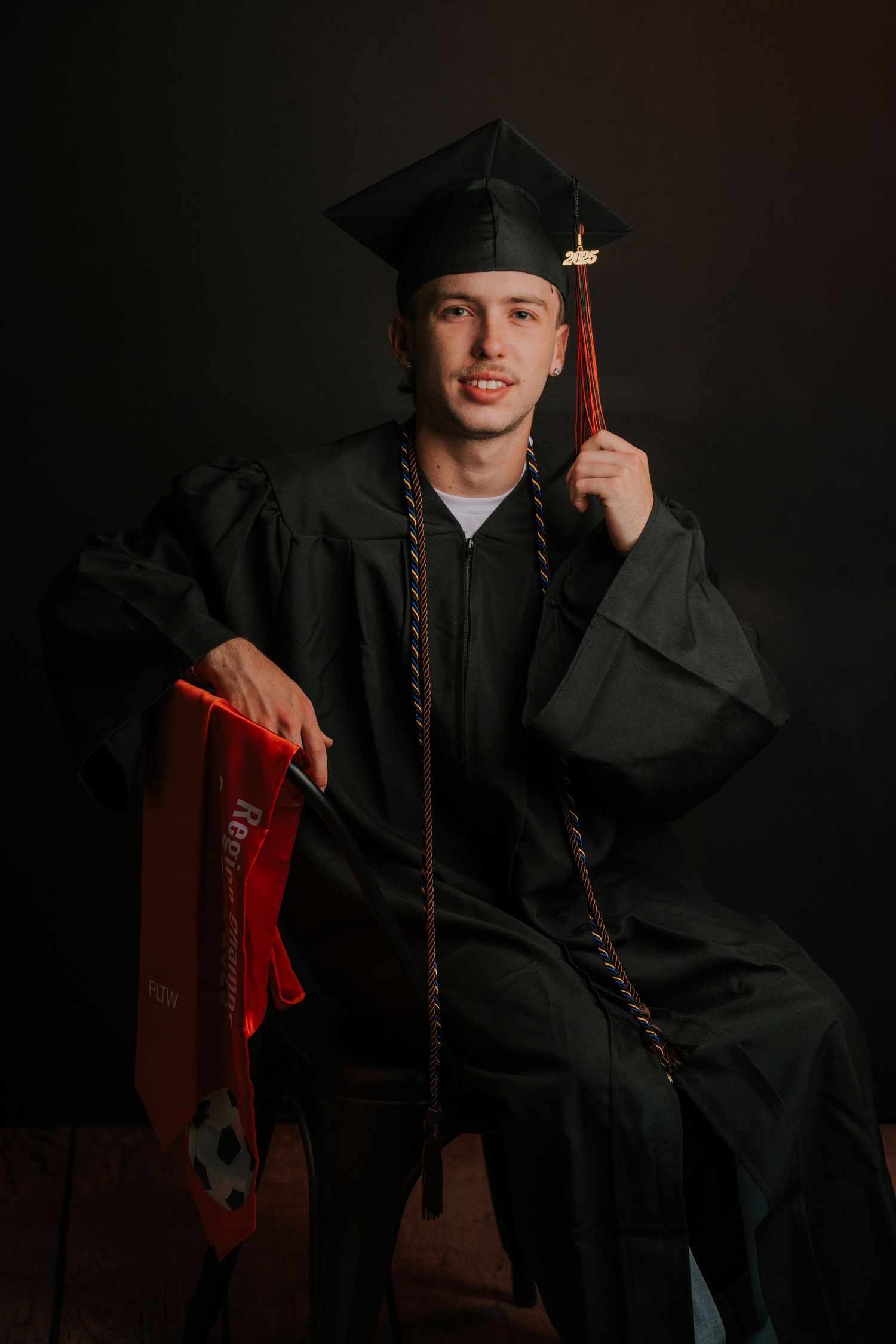 Graduate in traditional black cap and gown poses with orange honors cords.