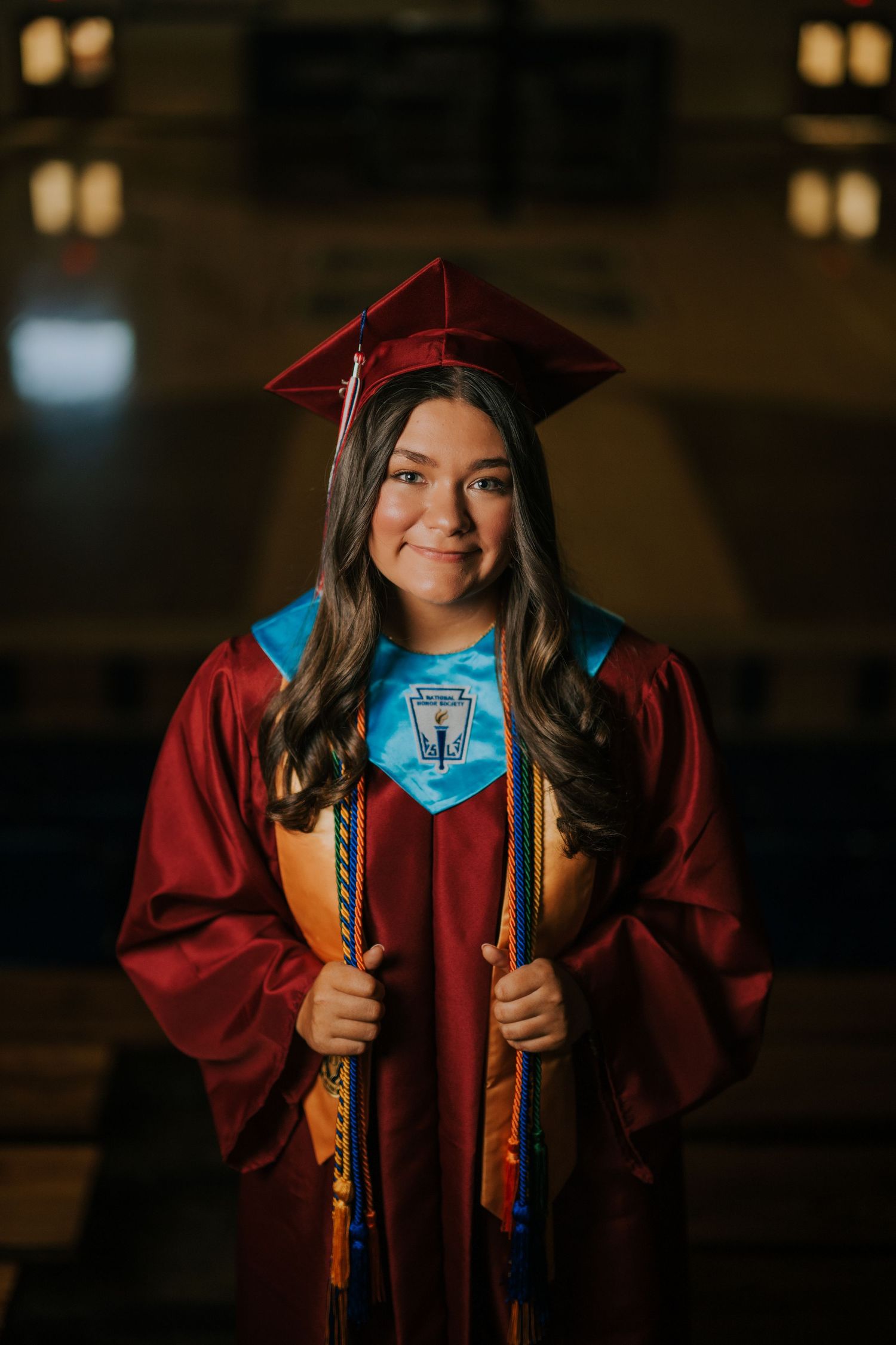 Graduate in red cap and gown with blue stole and honor cords posing indoors.