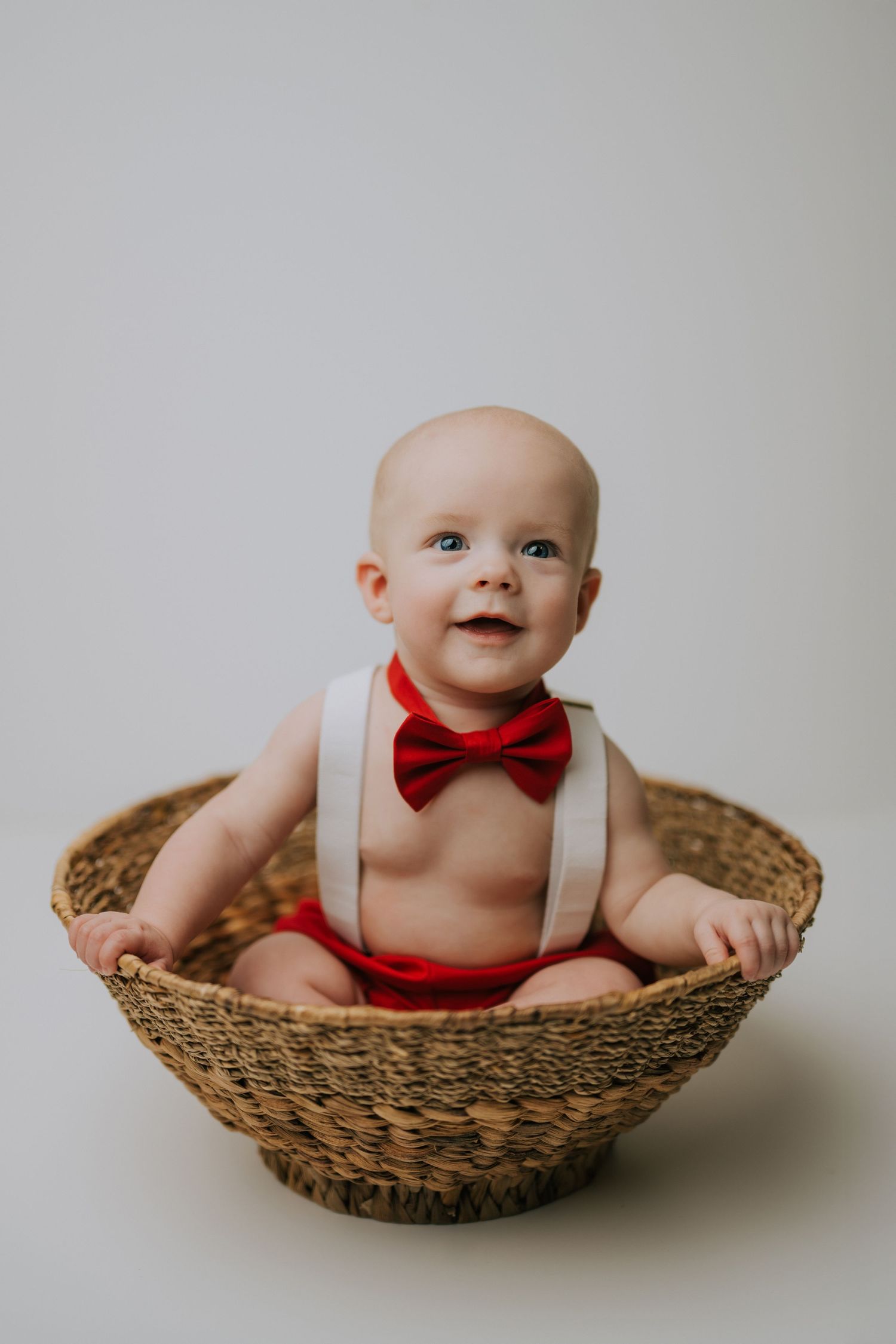 Baby wearing red bow tie and suspenders sits smiling in woven basket against white background.