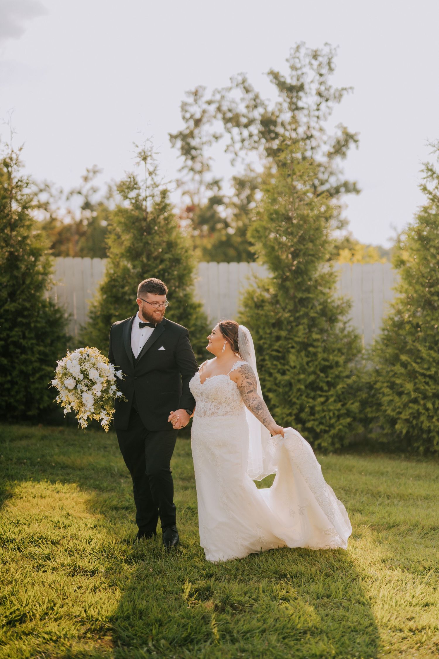 A couple walks hand in hand on their wedding day across a sunlit lawn with evergreen trees.