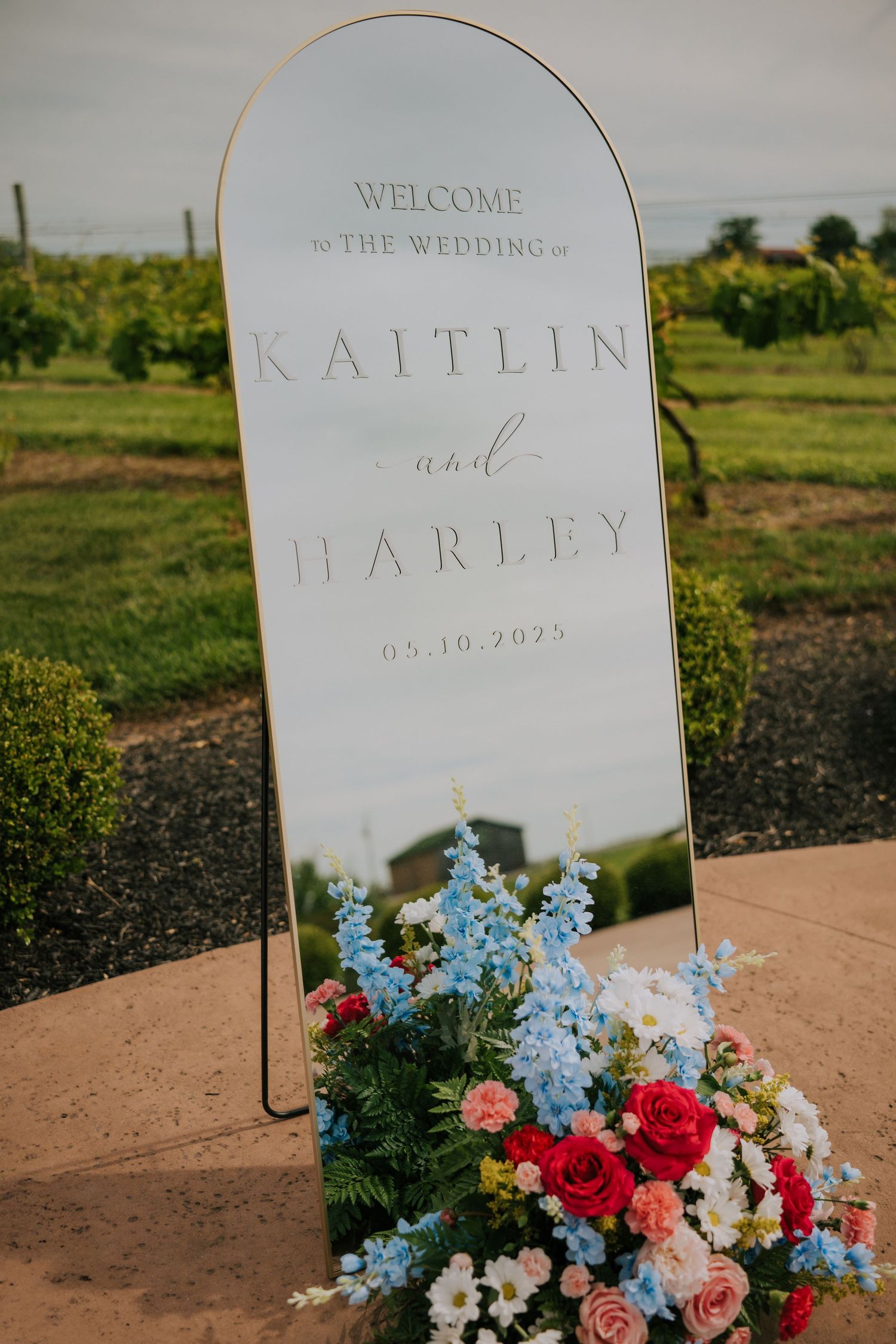 Elegant wedding welcome sign with floral arrangements in blue and red displayed on a curved mirror stand.