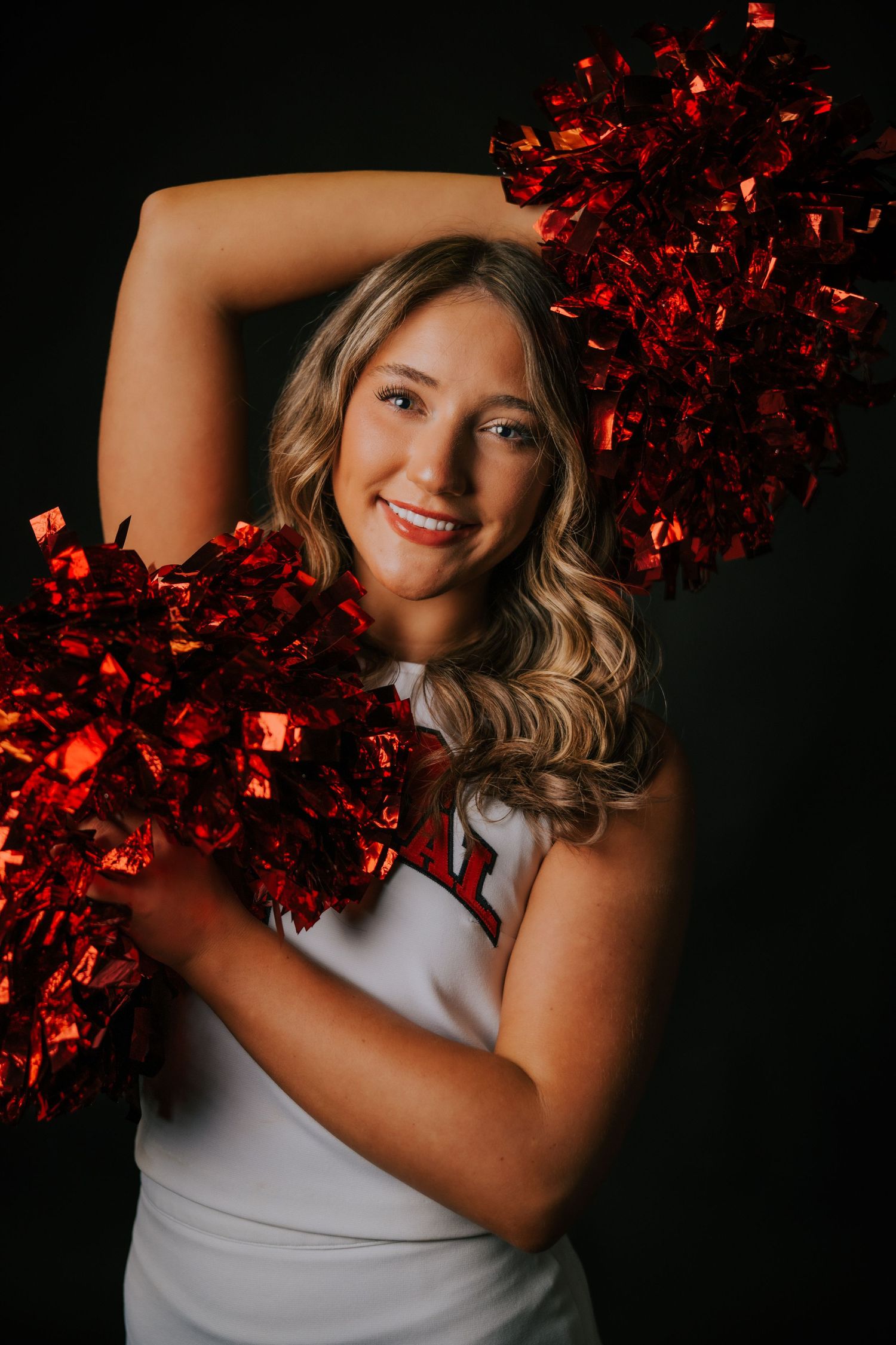 A figure in a white tank top poses playfully with red metallic pom poms against a dark background.