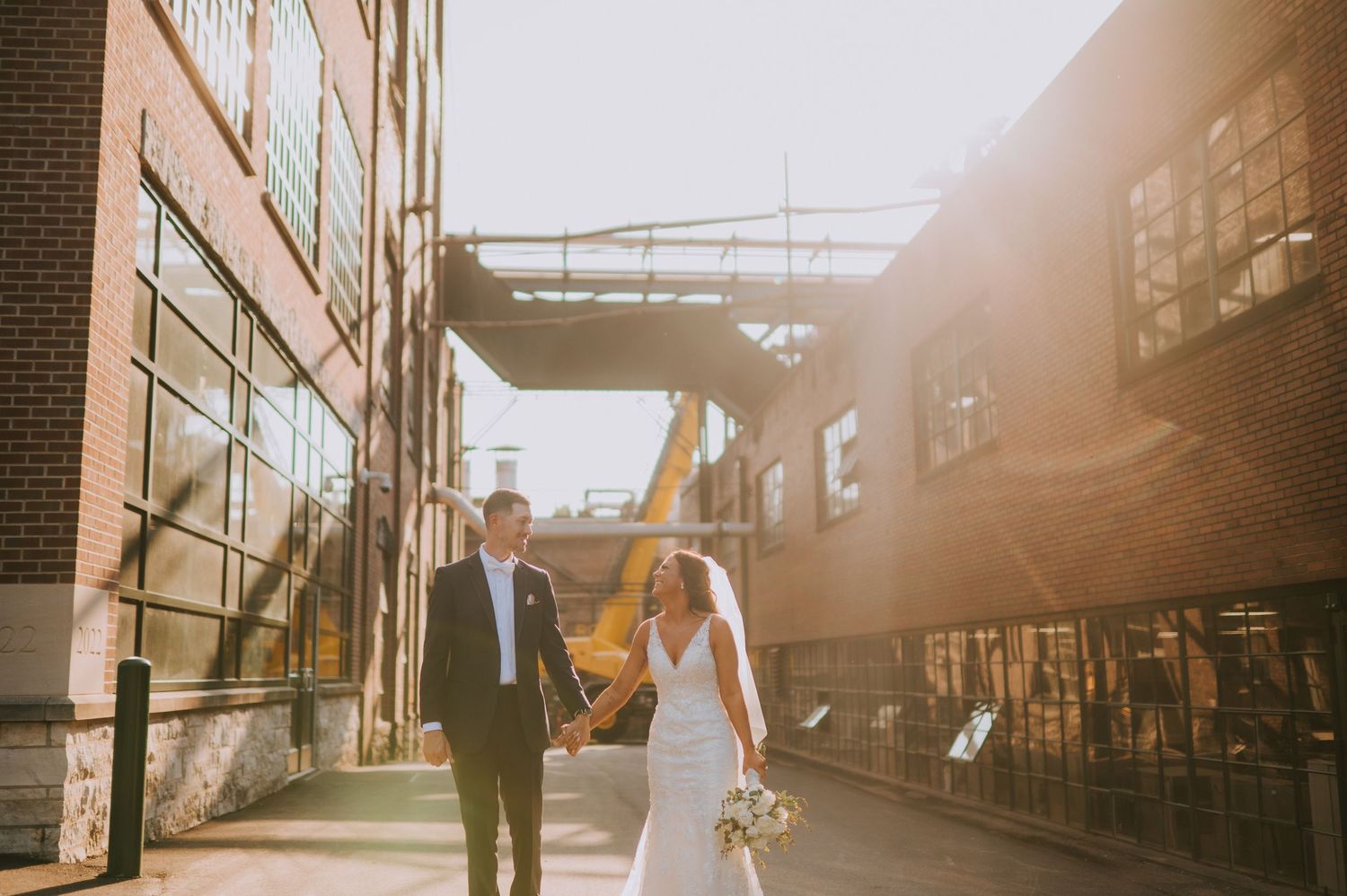 Newlyweds walking hand in hand through industrial building complex at sunset.