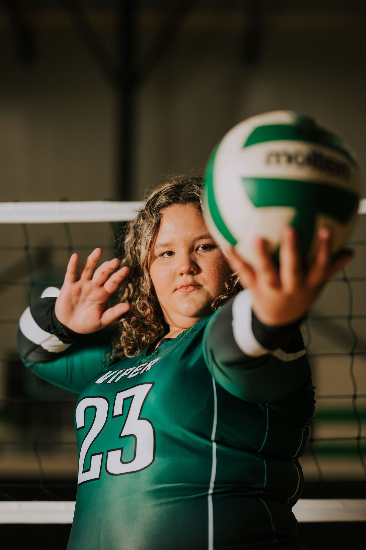 Volleyball player in green jersey number 23 poses defensively at the net with a green and white volleyball.