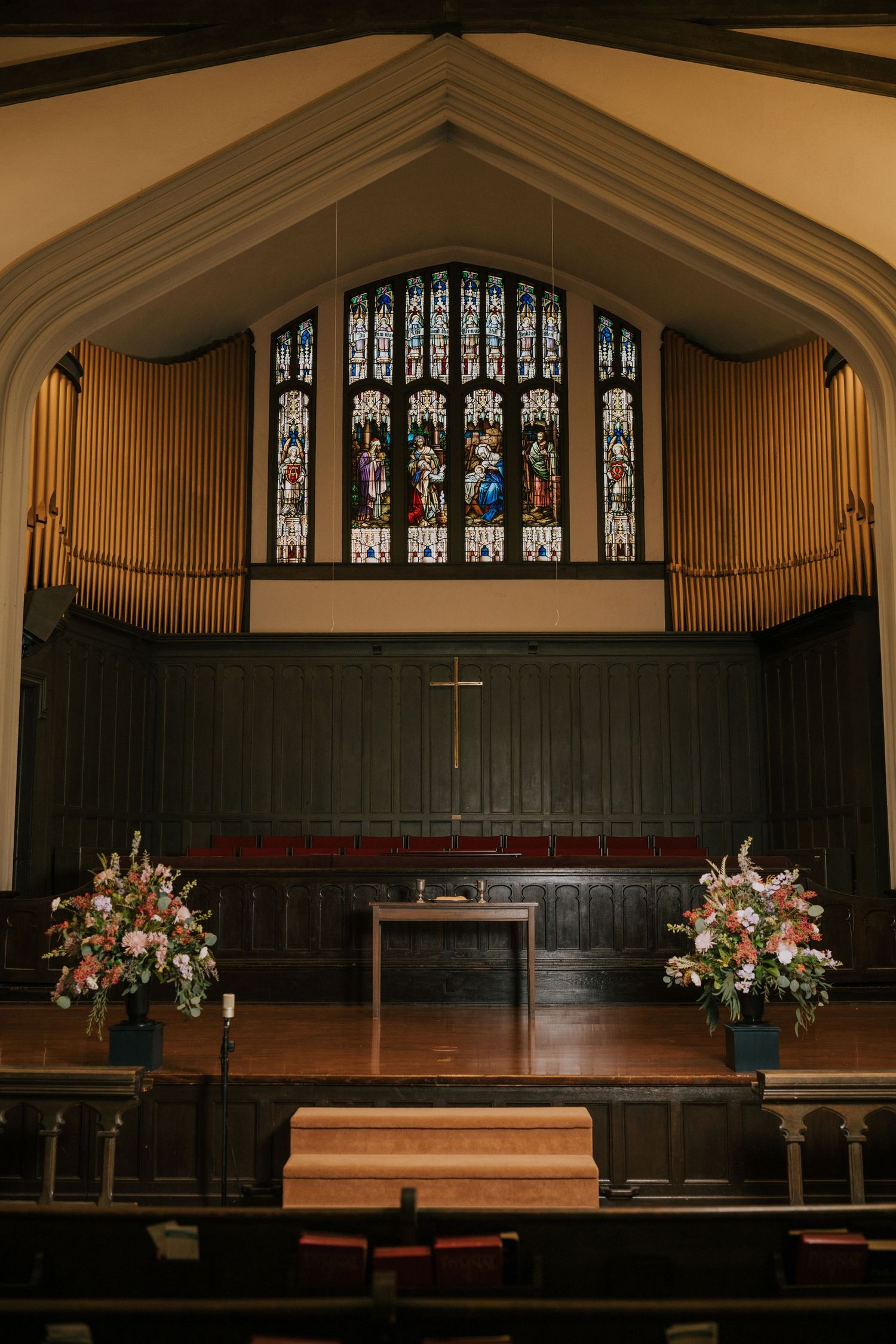 Church interior with stained glass window, organ pipes, wooden altar and floral arrangements in a traditional sanctuary.
