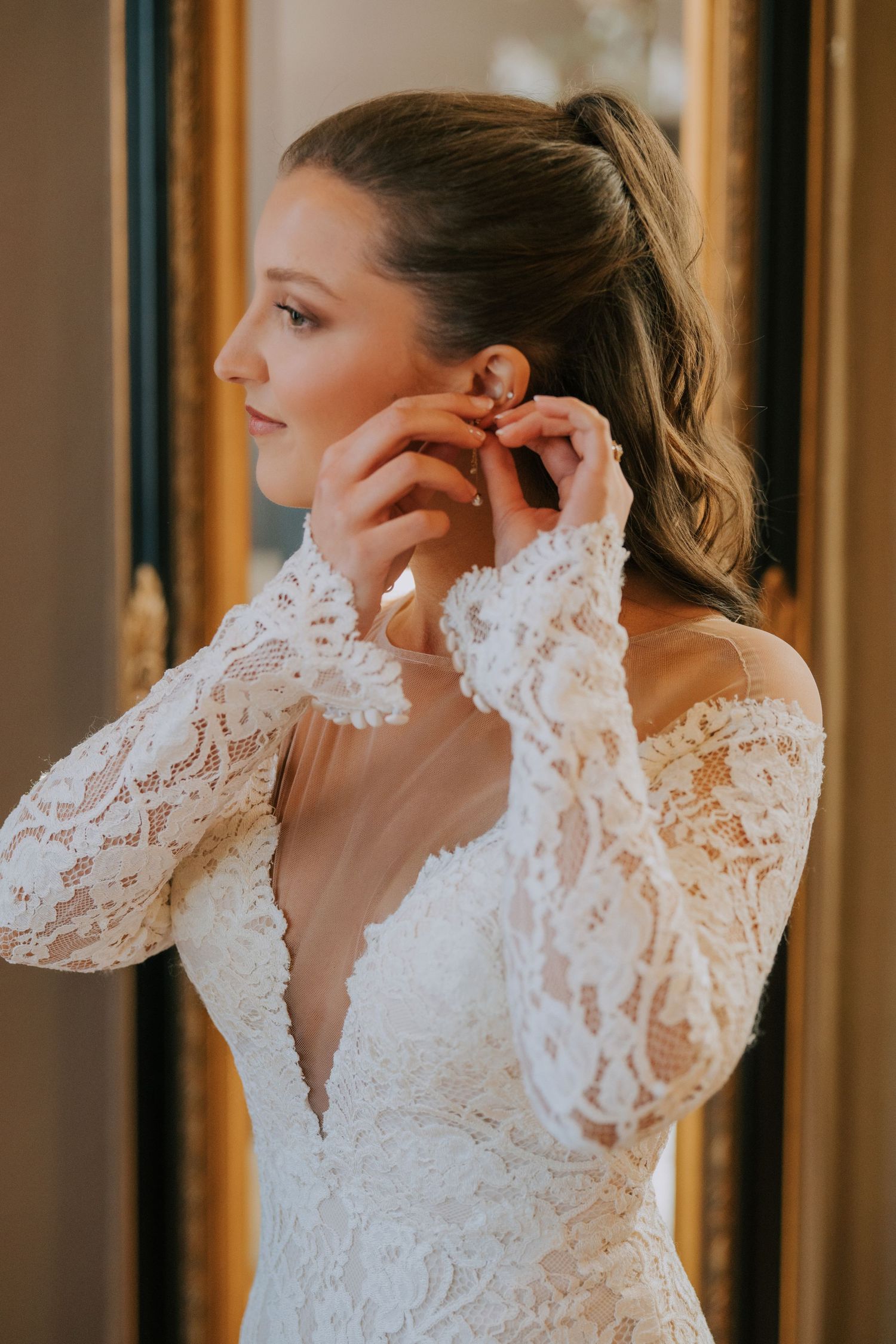 Side profile of a bride in a long-sleeved lace wedding dress putting on earrings in golden sunlight.