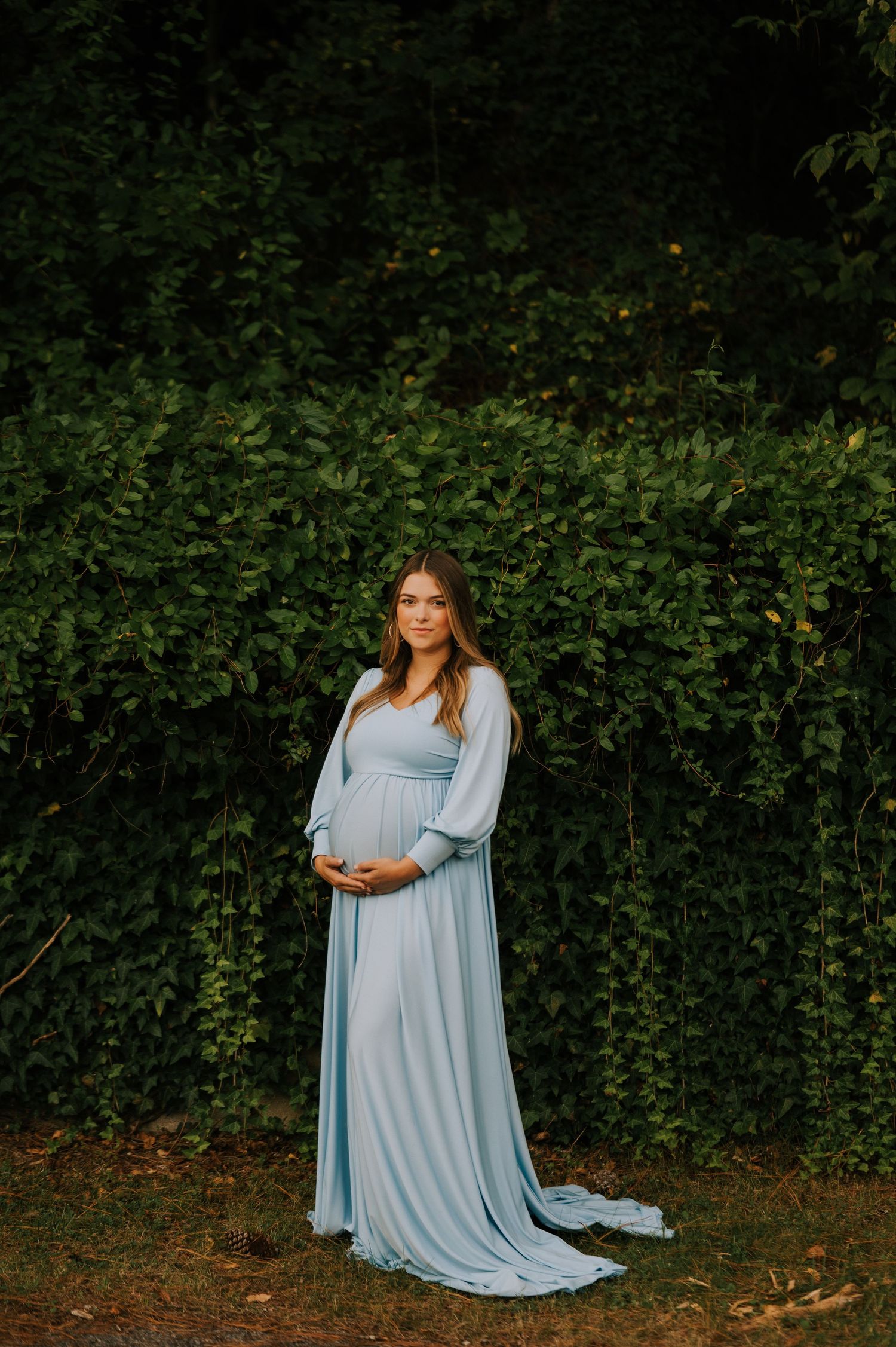 A maternity portrait with a woman wearing a pale blue gown against lush greenery.