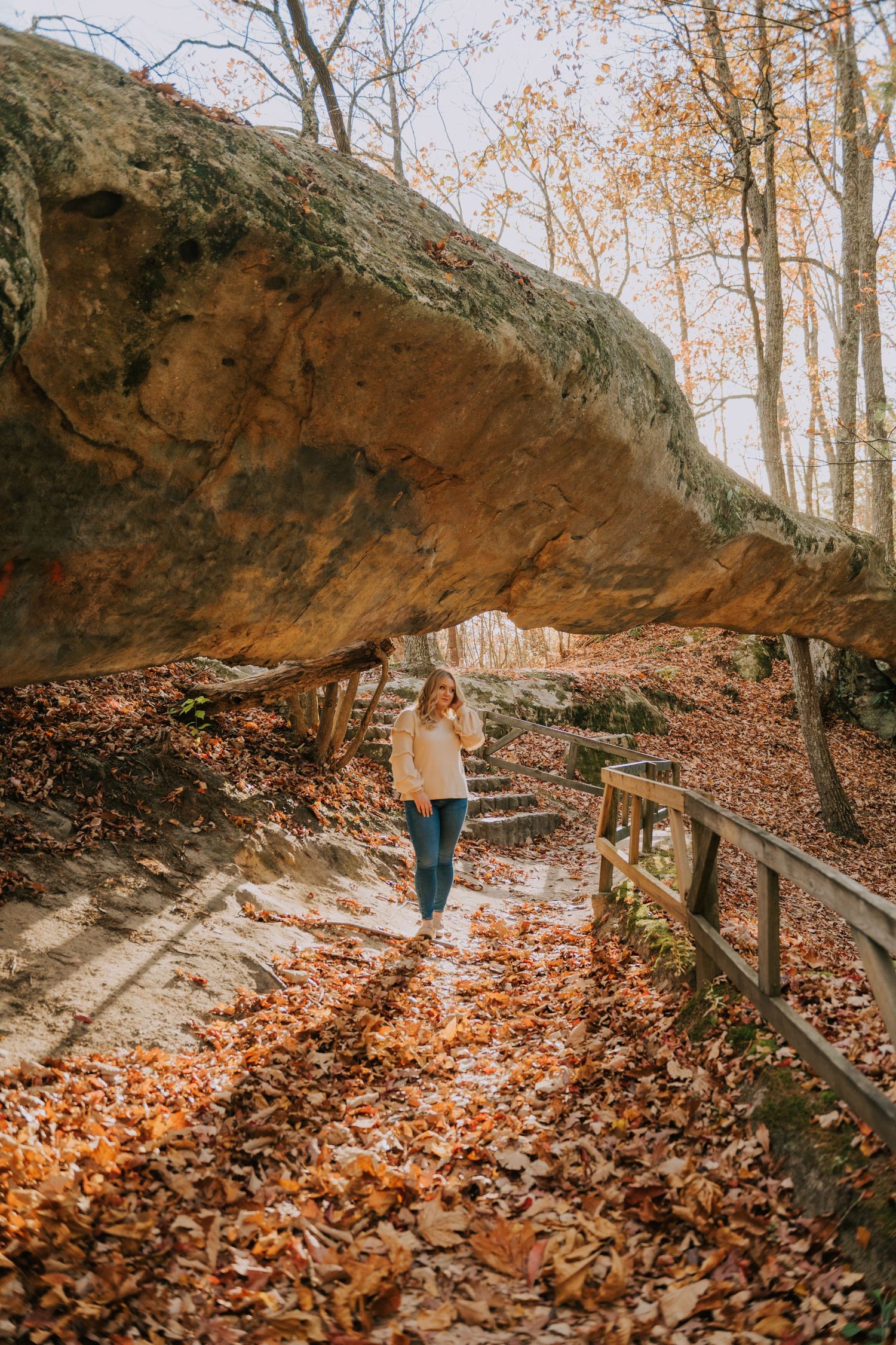 A scenic autumn trail leads beneath a large natural rock overhang with fallen leaves covering the ground.