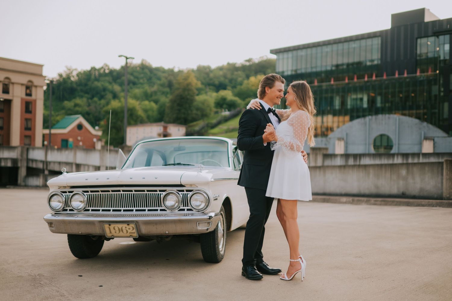 A couple poses near vintage cars on a city street with classic buildings in the background.