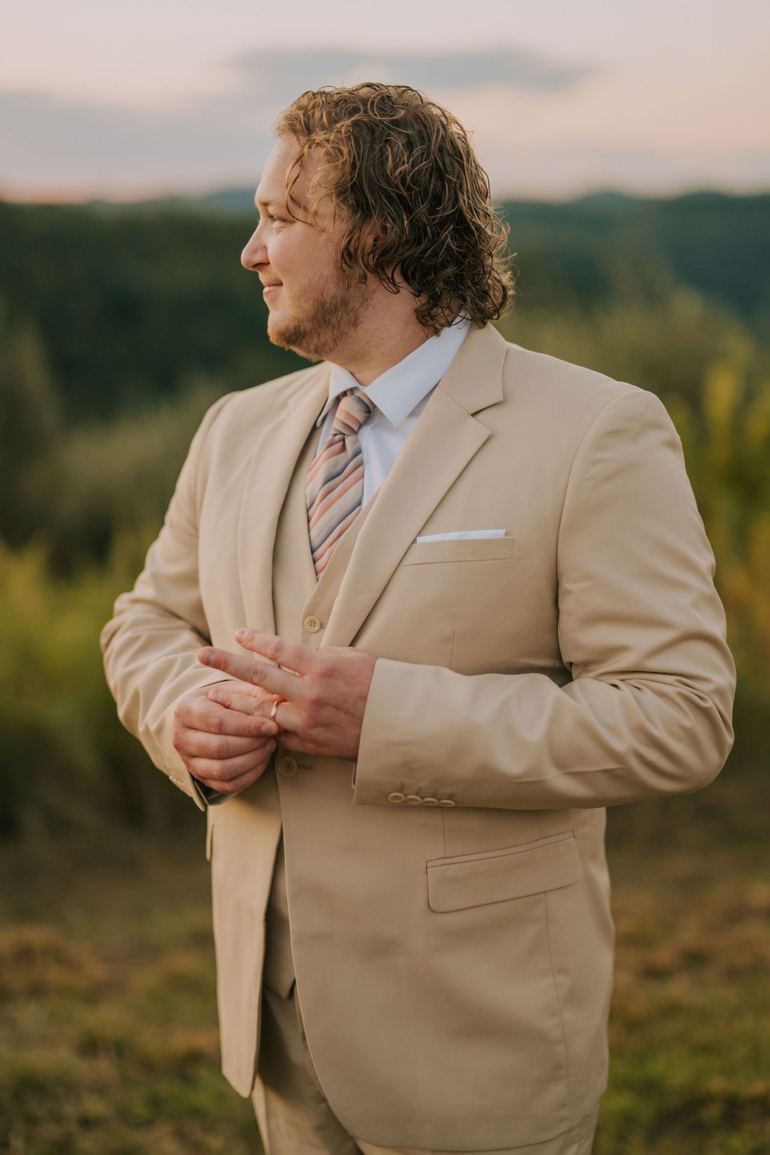Groom in tan suit adjusting his jacket against mountain backdrop.