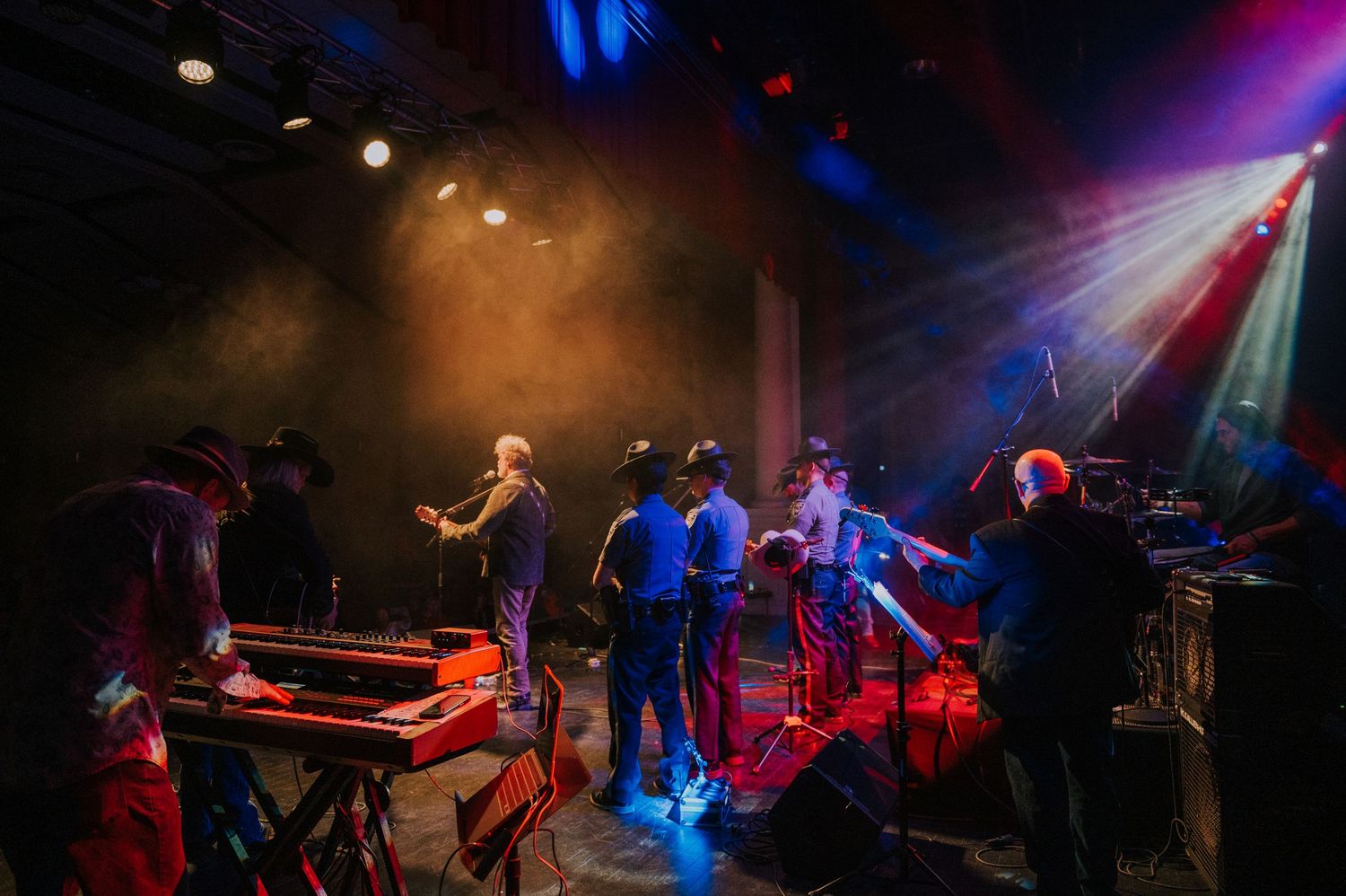A band performs on a dimly lit stage with dramatic colorful lighting and smoke effects during a live concert performance.