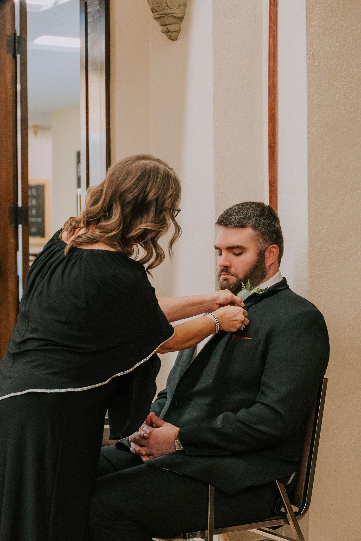 A person helps adjust formal attire of someone seated in a chair during wedding preparations.