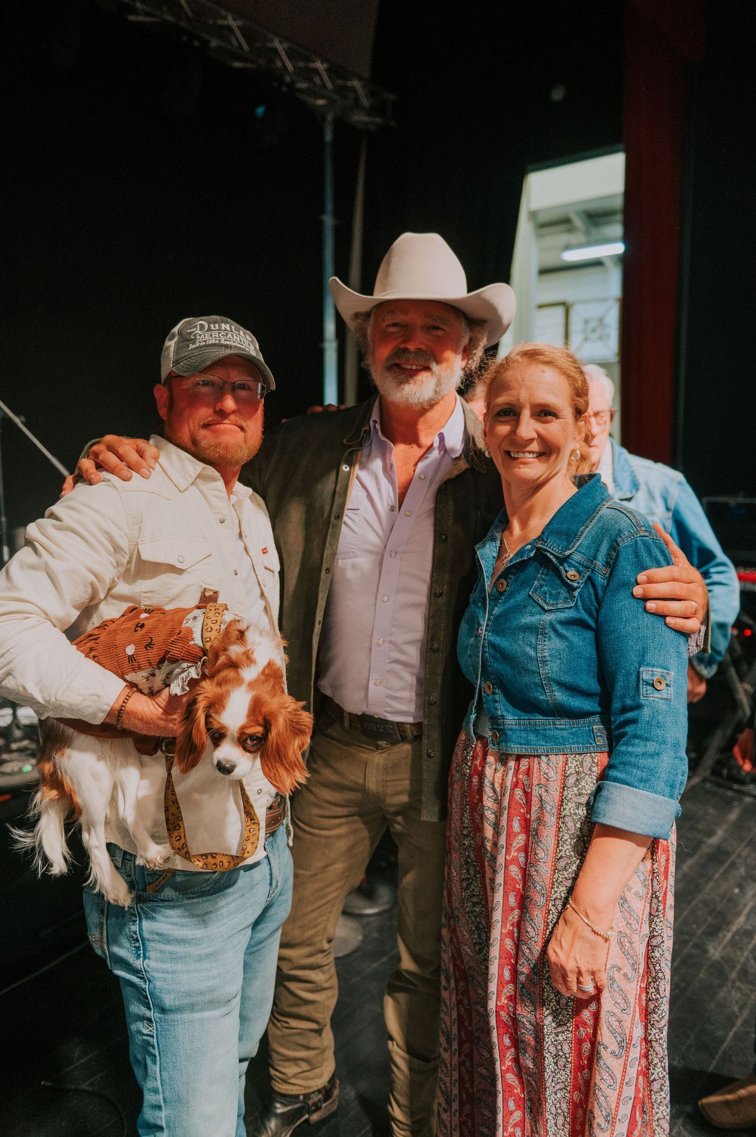 Three people wearing casual western attire pose together backstage at an event venue.