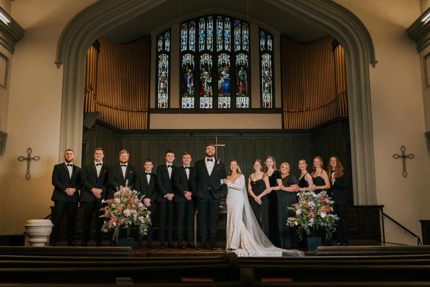 A wedding party stands arranged on church altar steps with stained glass windows and floral arrangements in the background.