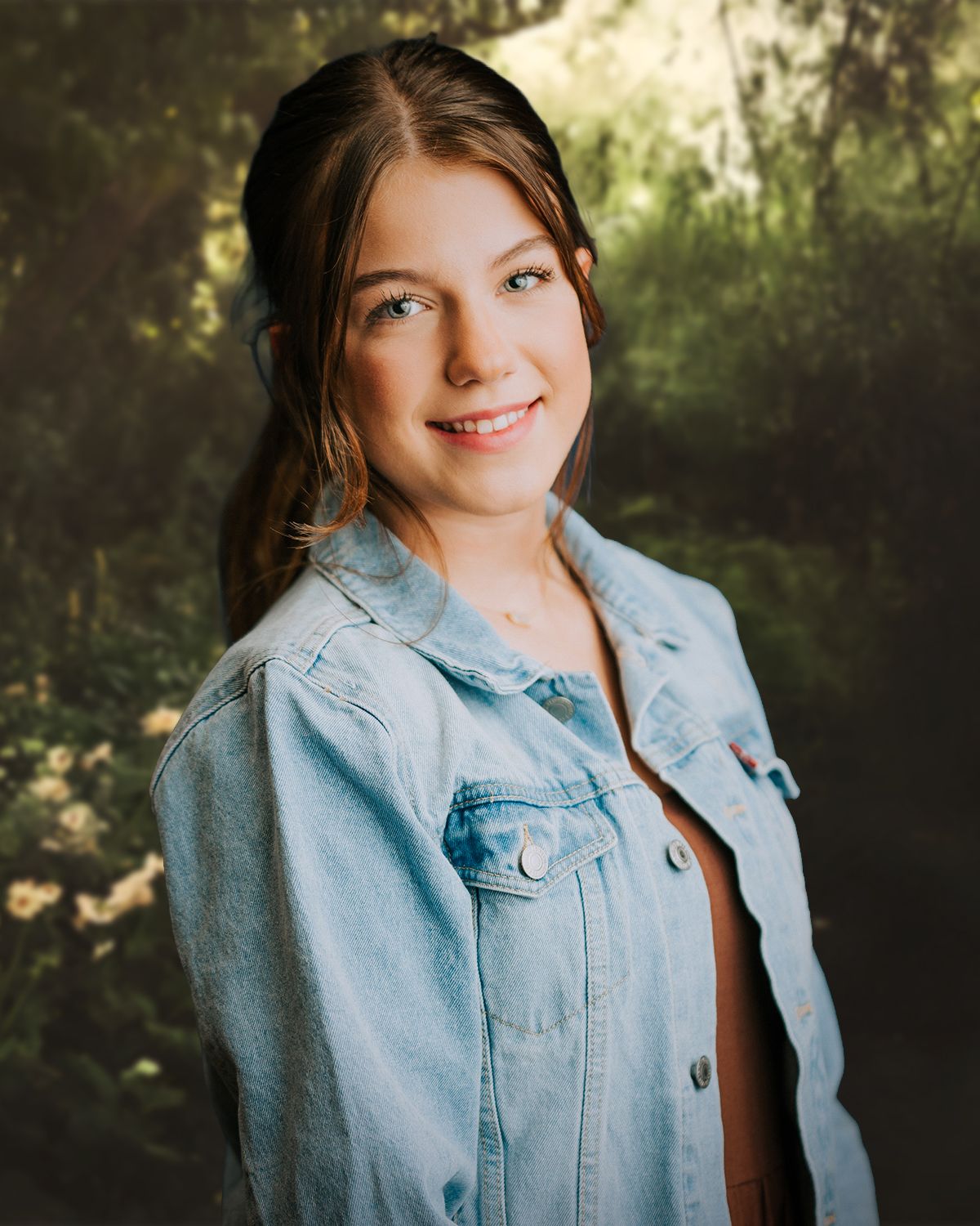 A smiling young woman with brown hair wearing a denim jacket poses outdoors with a lush green background.