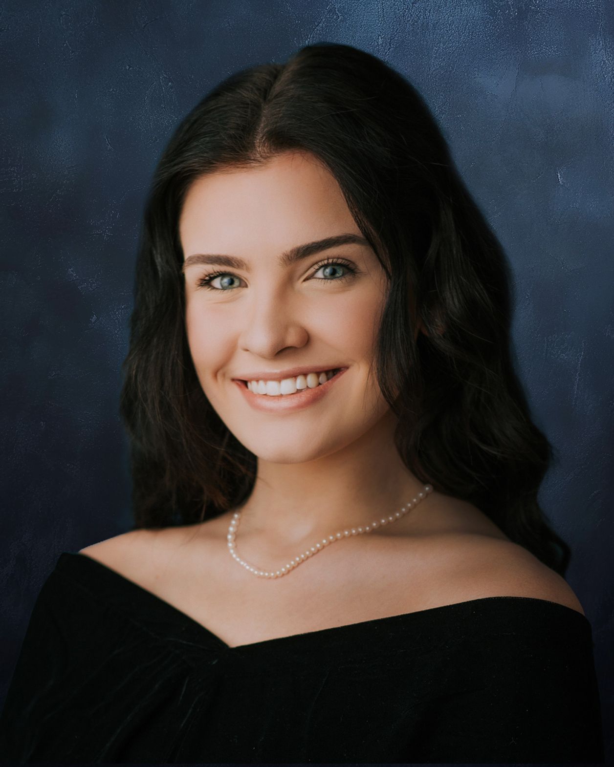 Young woman with dark hair wearing a black off-shoulder top and pearl necklace, smiling in a formal portrait.