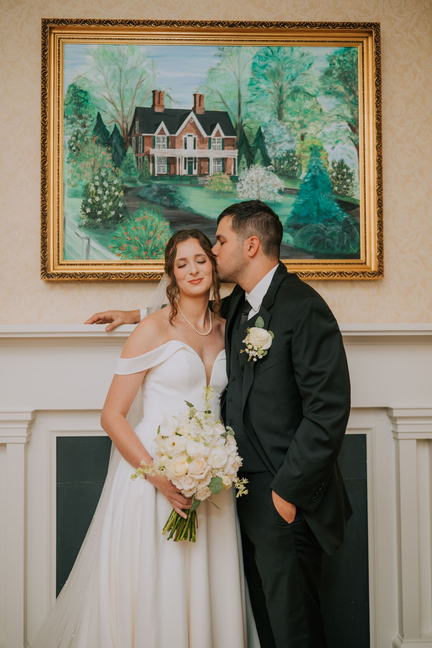 A romantic wedding couple embraces in front of framed landscape paintings and white wainscoting at their venue.
