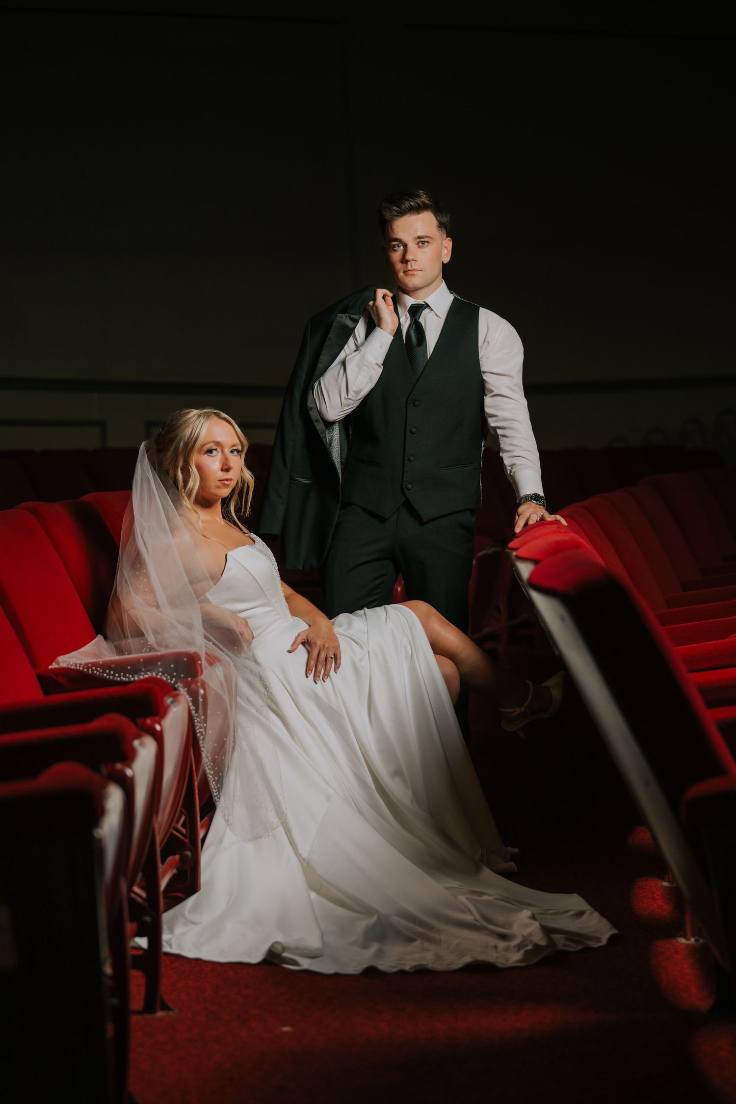 Dramatic wedding portrait in dark theater with couple seated on red velvet chairs in moody lighting.