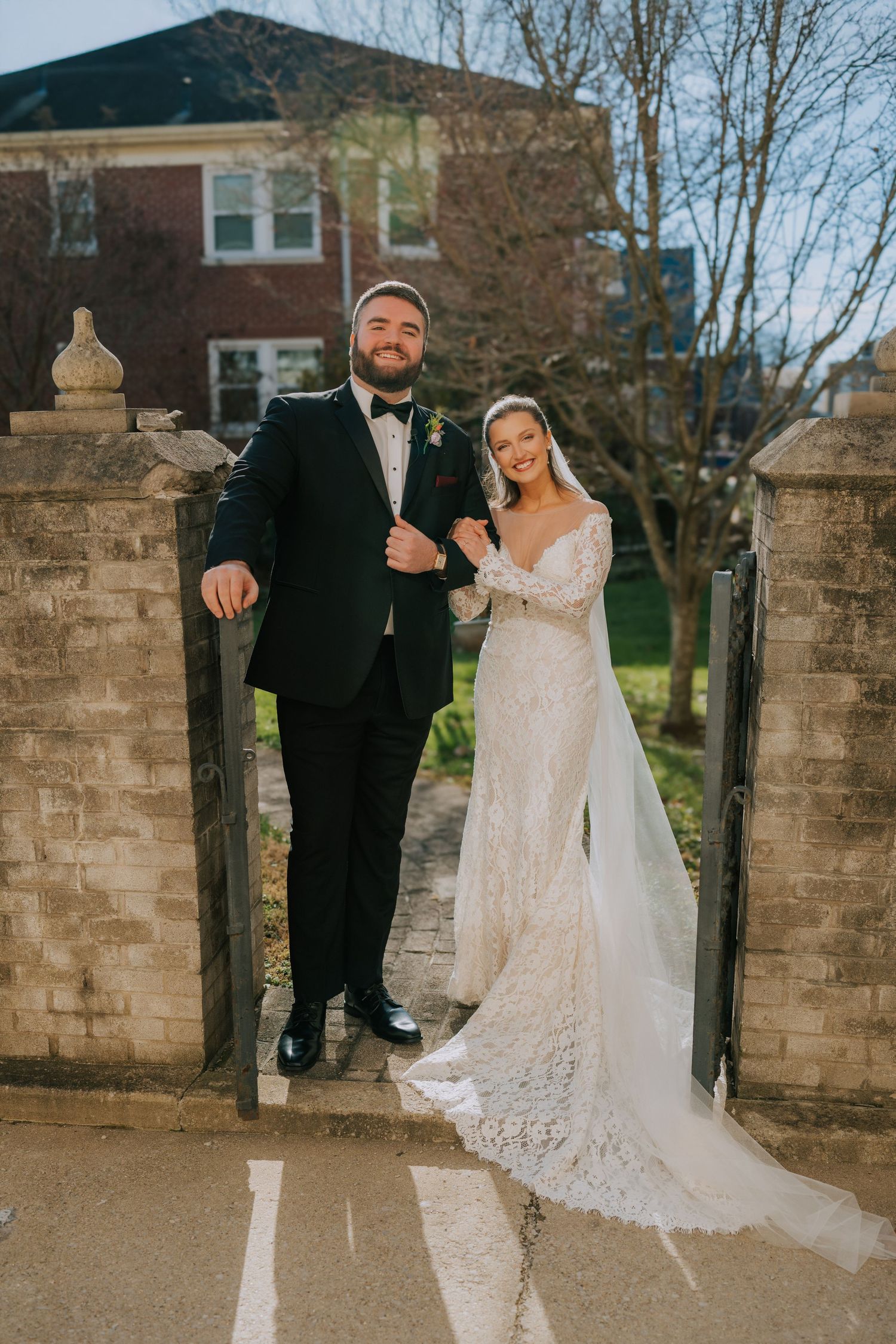 Two people in formal wedding attire pose between stone pillars on a flagstone path outside a brick building.