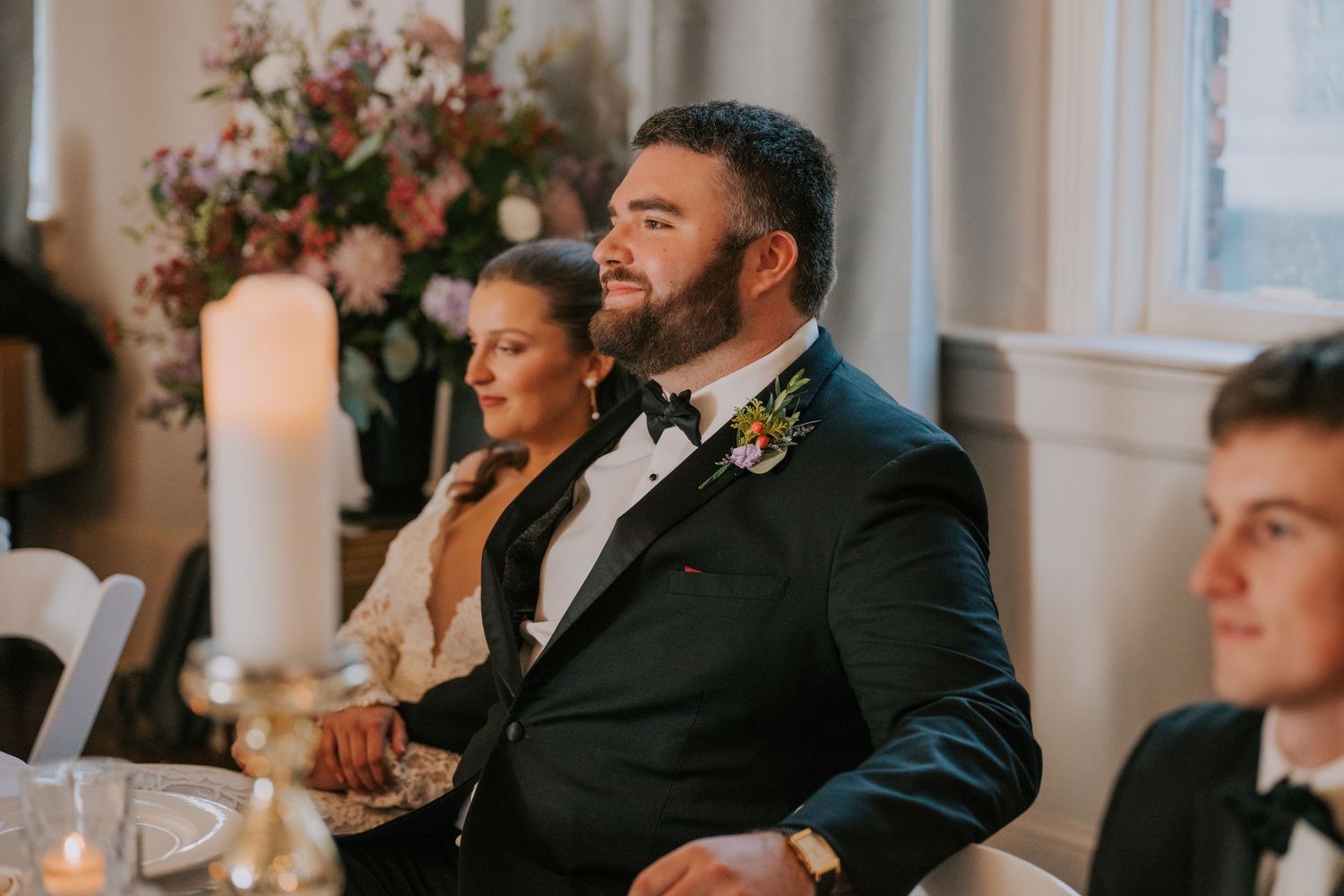 Wedding guests enjoy an elegant reception dinner with floral arrangements and candlelight at a formal indoor venue.