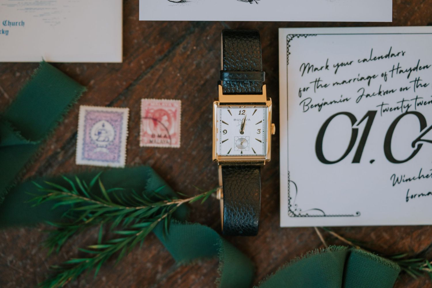 Vintage stamps and a classic wristwatch with green leather strap arranged on a rustic surface with greenery and a handwritten note.