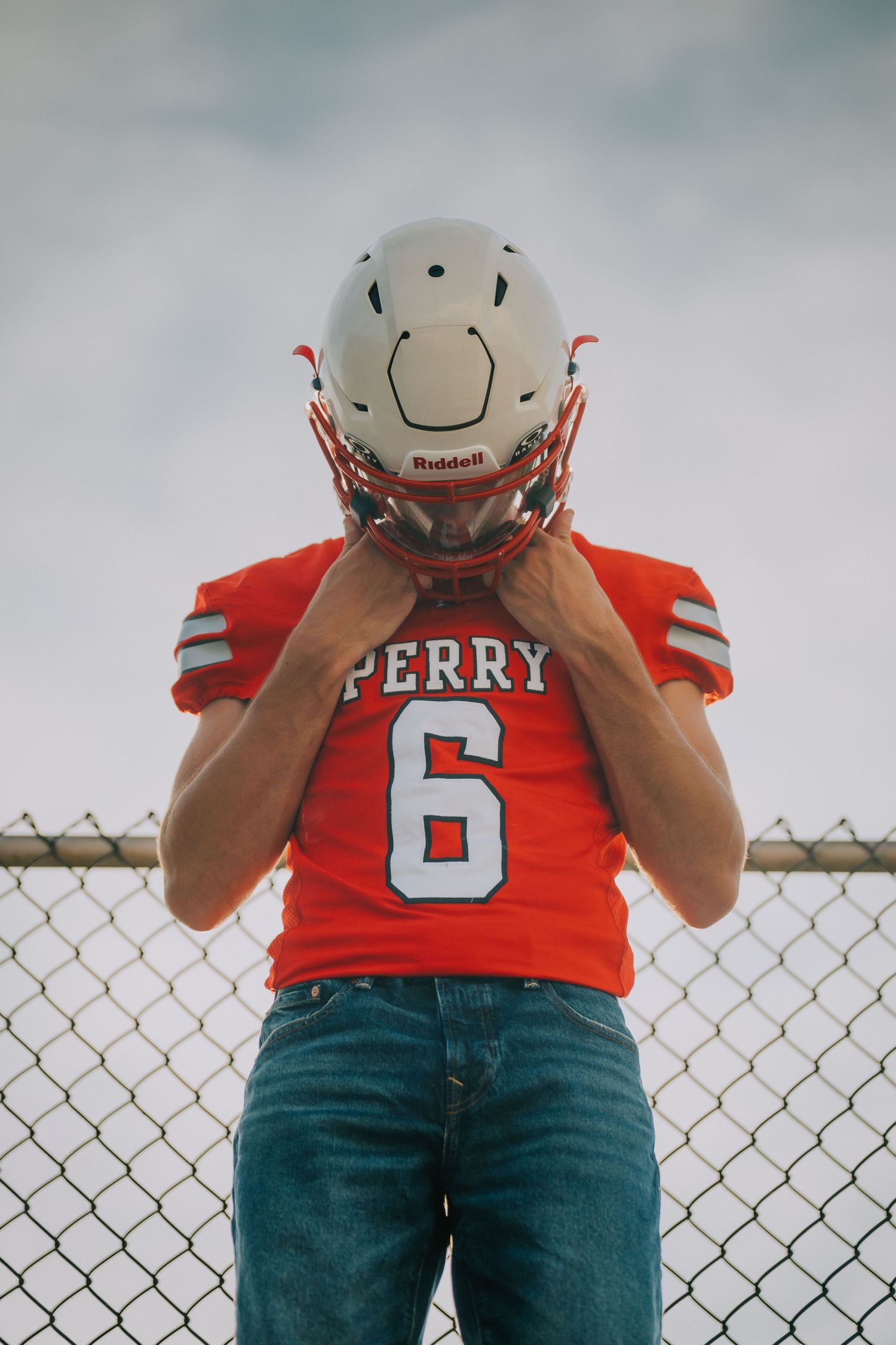A football player wearing a red Perry #6 jersey and Riddell helmet poses against a chain link fence.