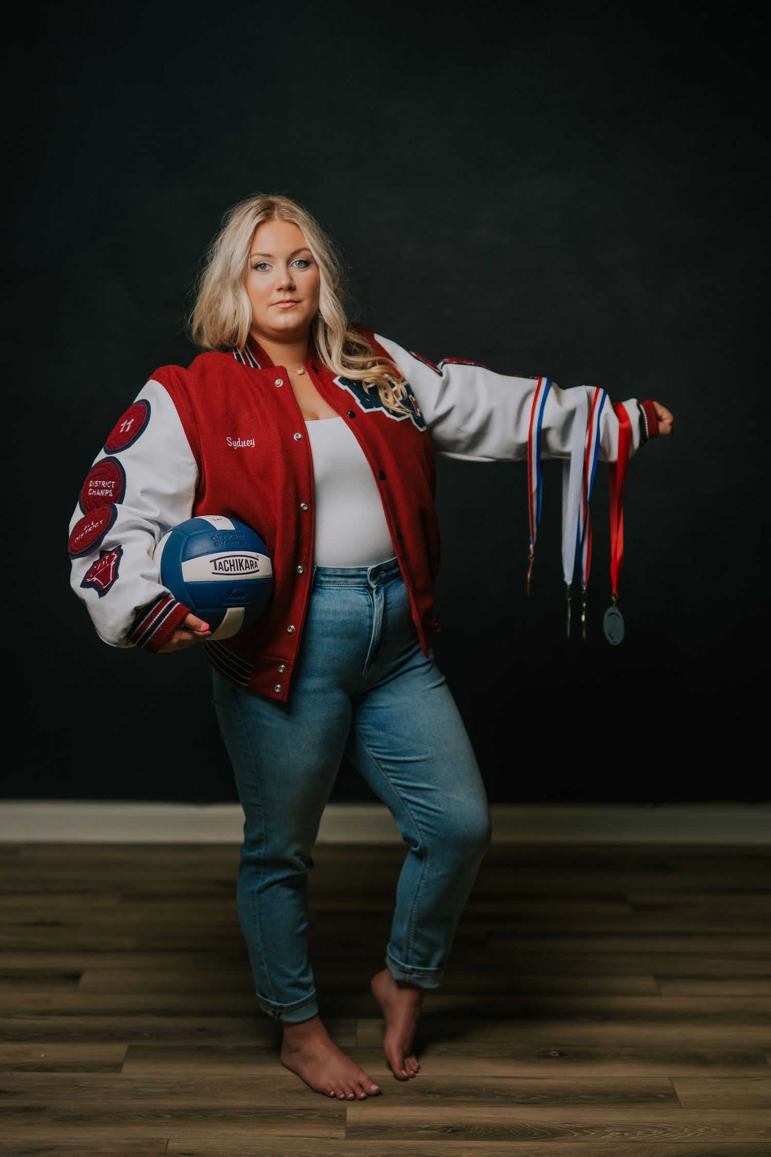Athletic varsity jacket and jeans outfit styled with barefoot pose against dark background holding sports equipment.