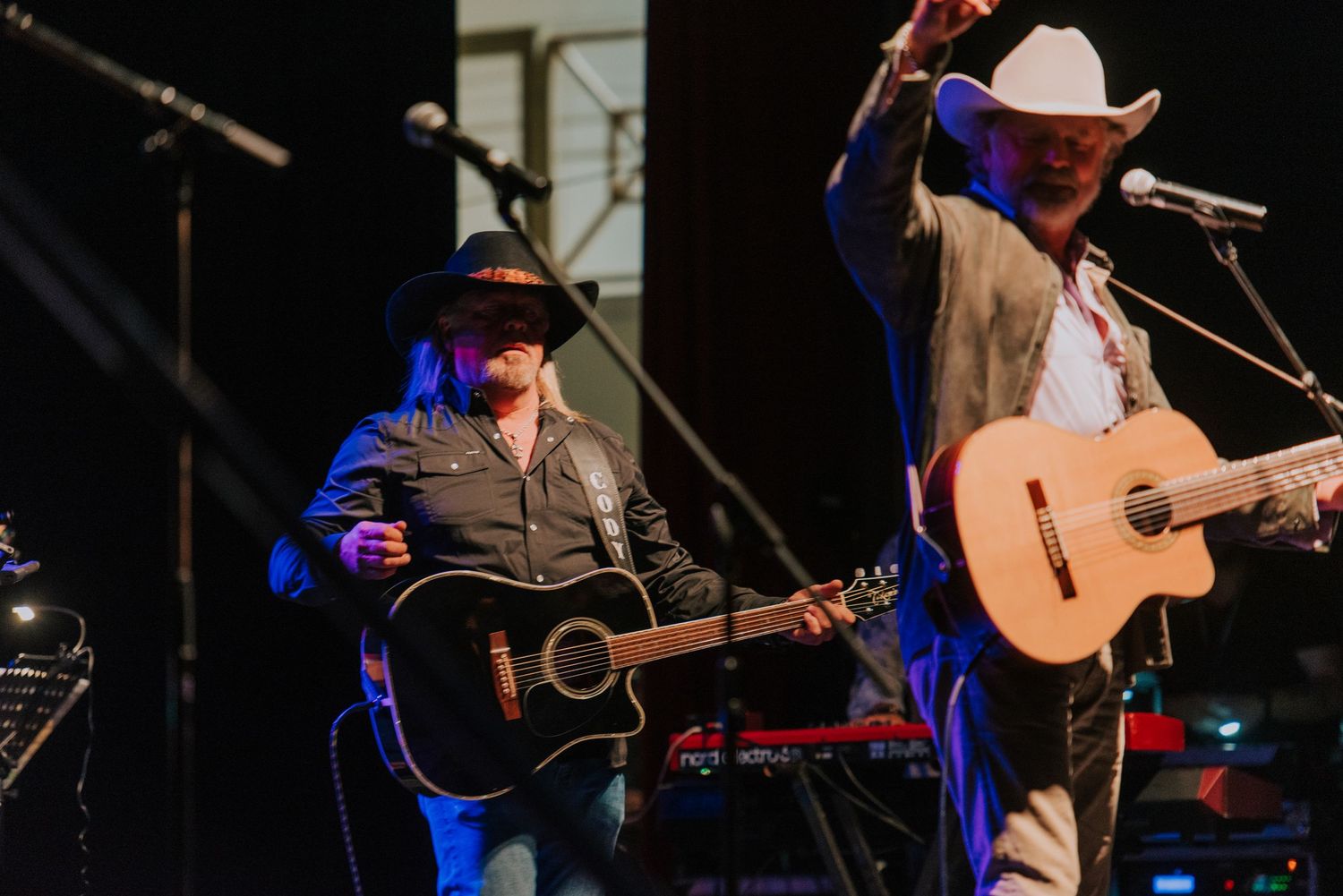 Two country western musicians perform with acoustic guitars under blue stage lighting at a concert venue.