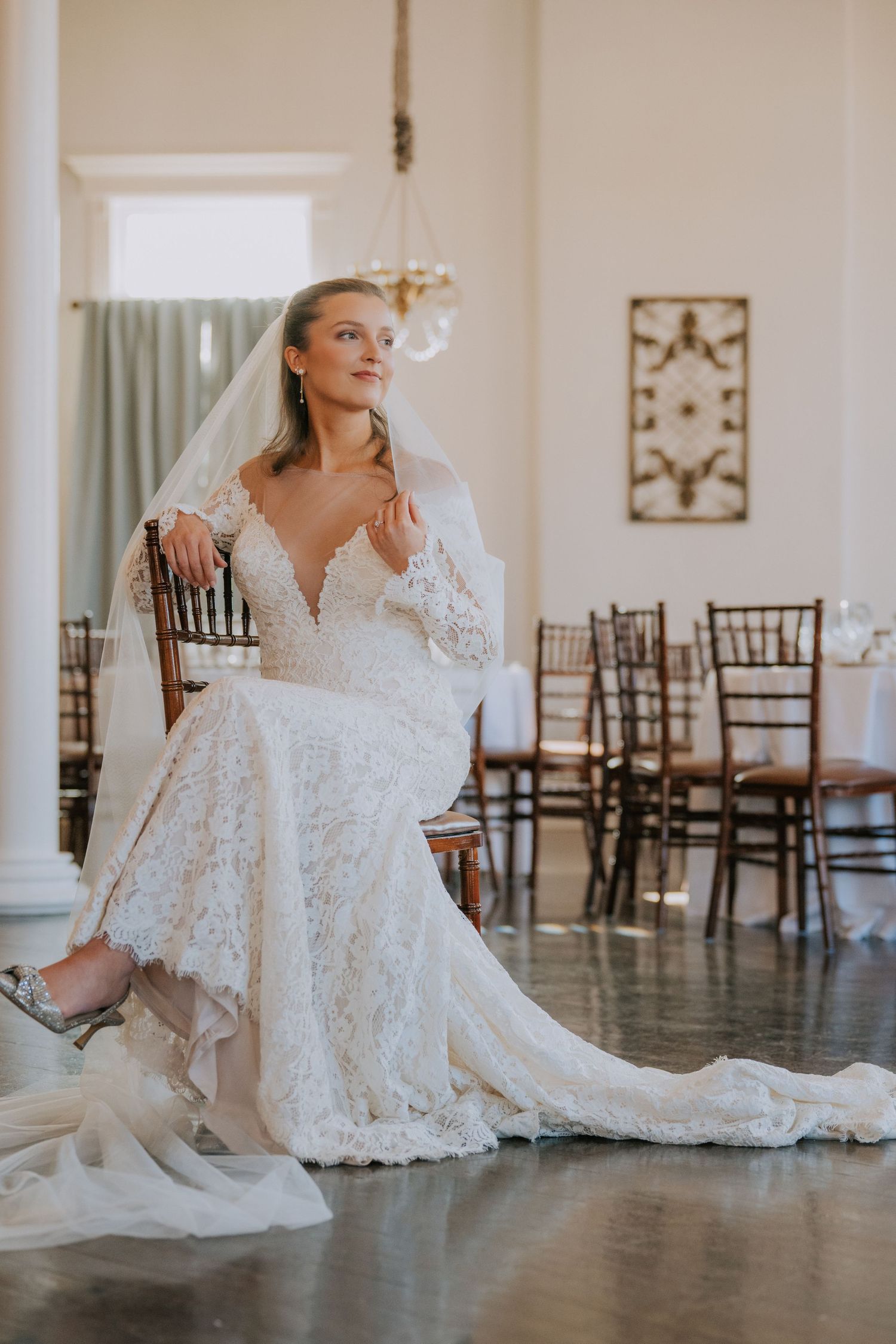 Bride in elegant lace wedding gown with plunging neckline sits gracefully on wooden chair in venue with empty chiavari chairs.