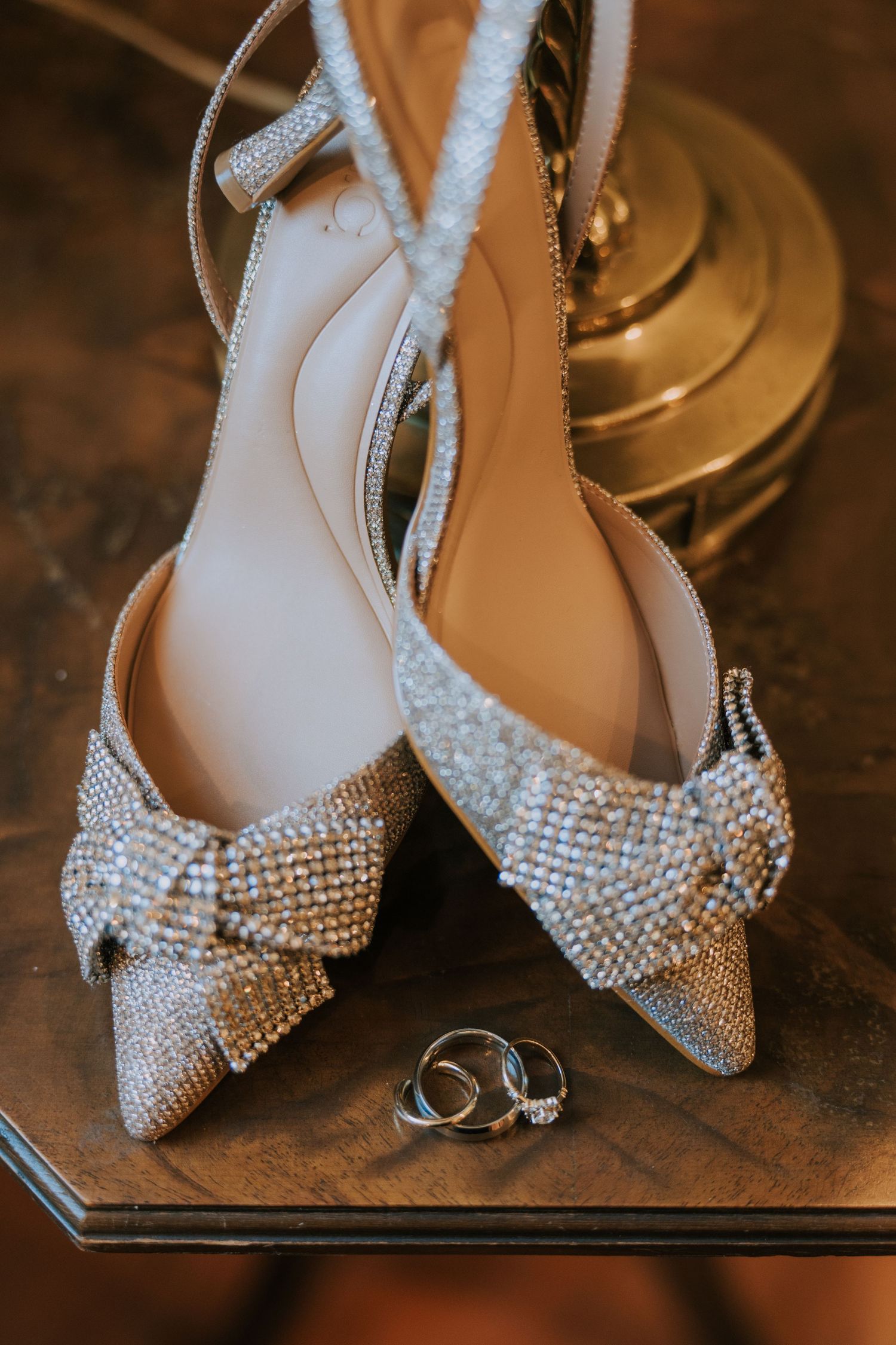 Sparkly rhinestone bridal shoes with bow detail and wedding rings displayed on wooden surface in warm lighting.