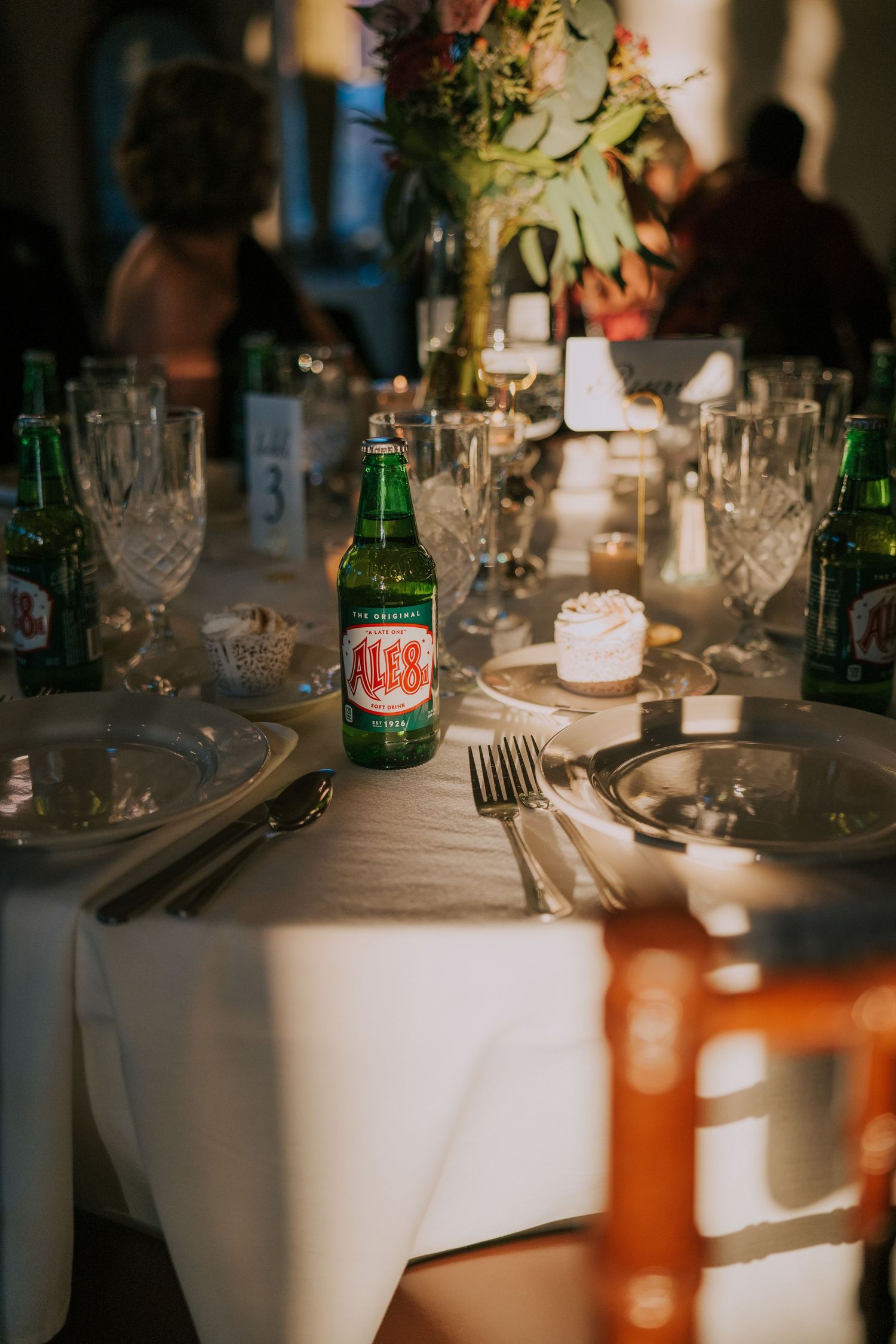 Wedding reception table settings with glassware and bottles under ambient lighting.