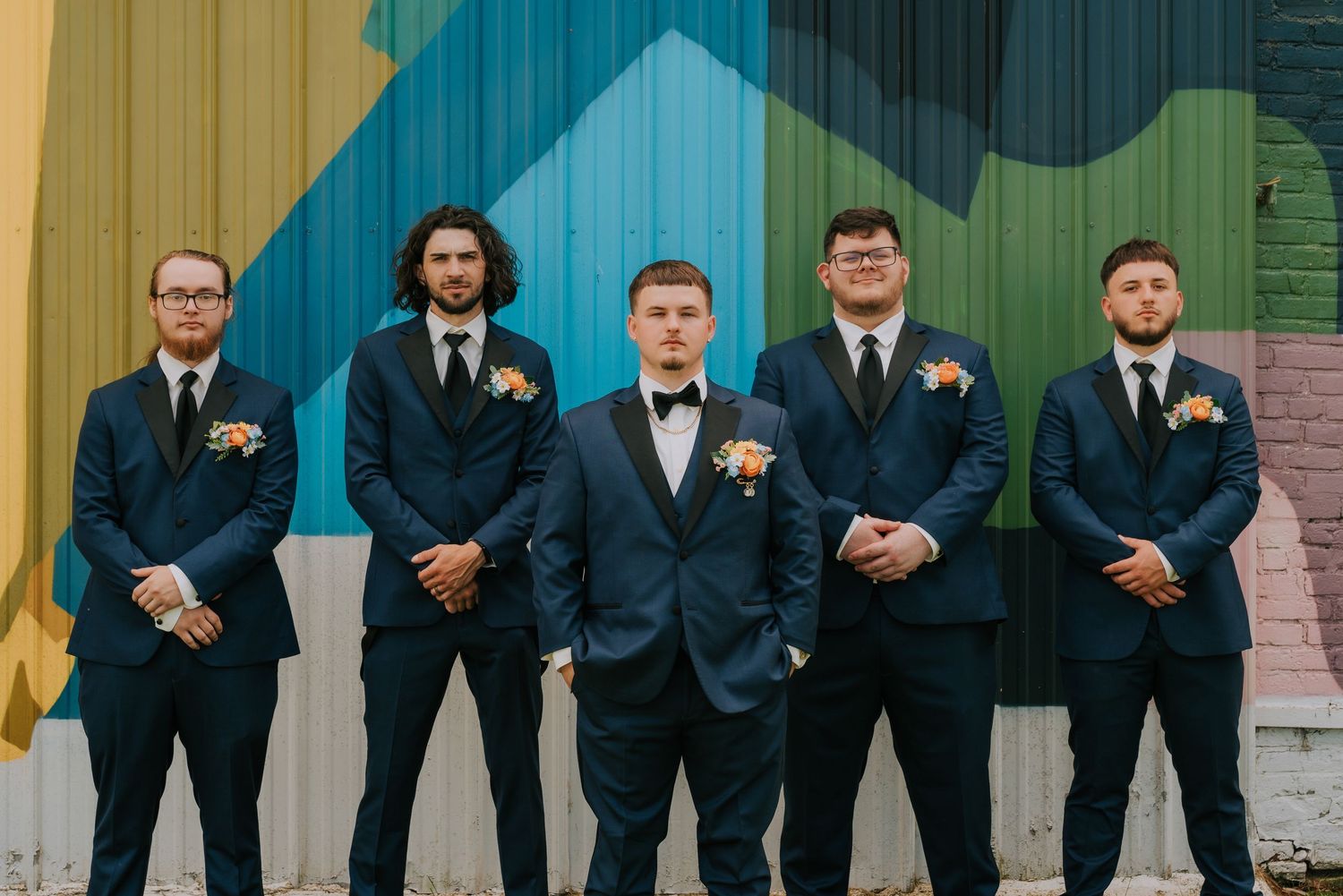 Groomsmen in dark suits lined up against colorful painted wall backdrop.