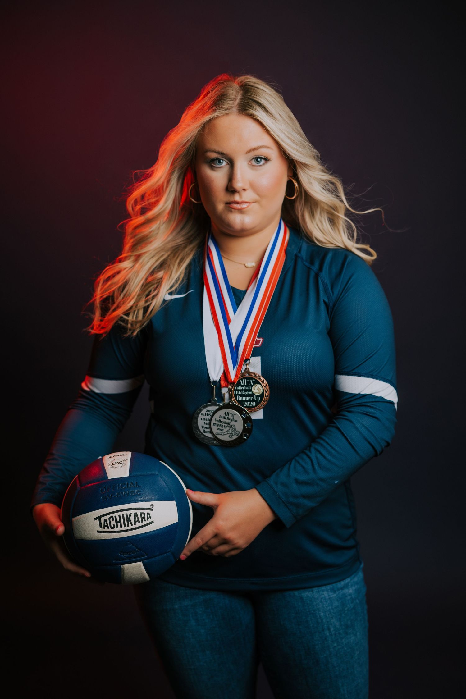 Student athlete poses with volleyball and medals against dark backdrop in dramatic sports portrait.