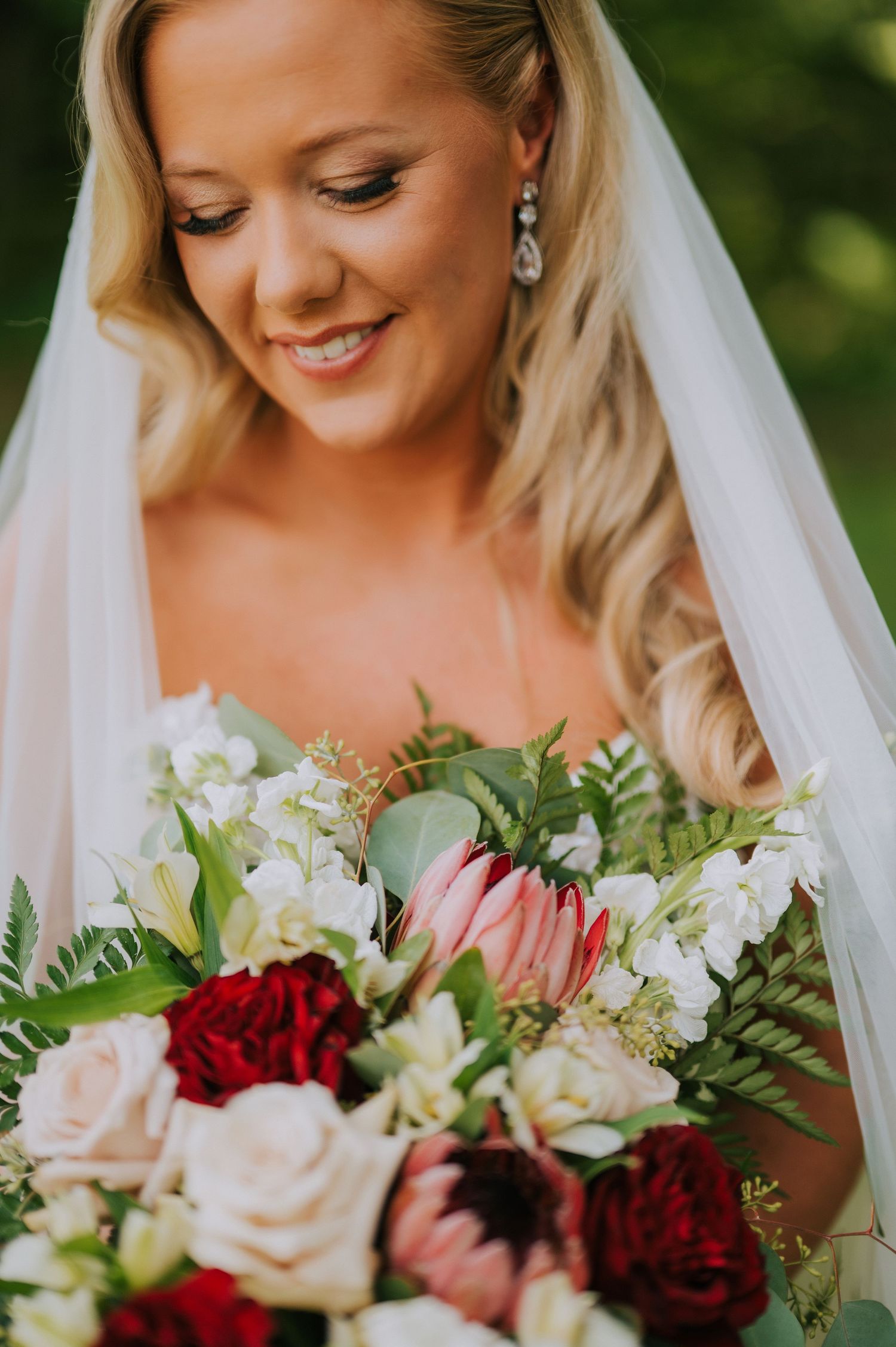 A bridal bouquet featuring deep red and blush roses with white accents and greenery against a soft outdoor background.