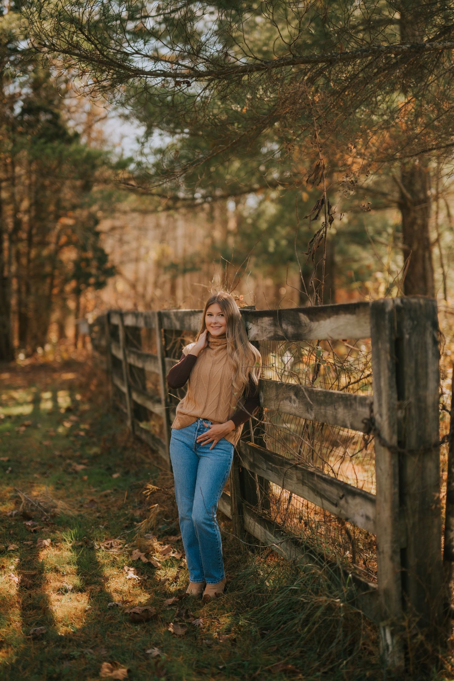 Autumn photoshoot by rustic wooden fence with golden sunlight filtering through pine trees.