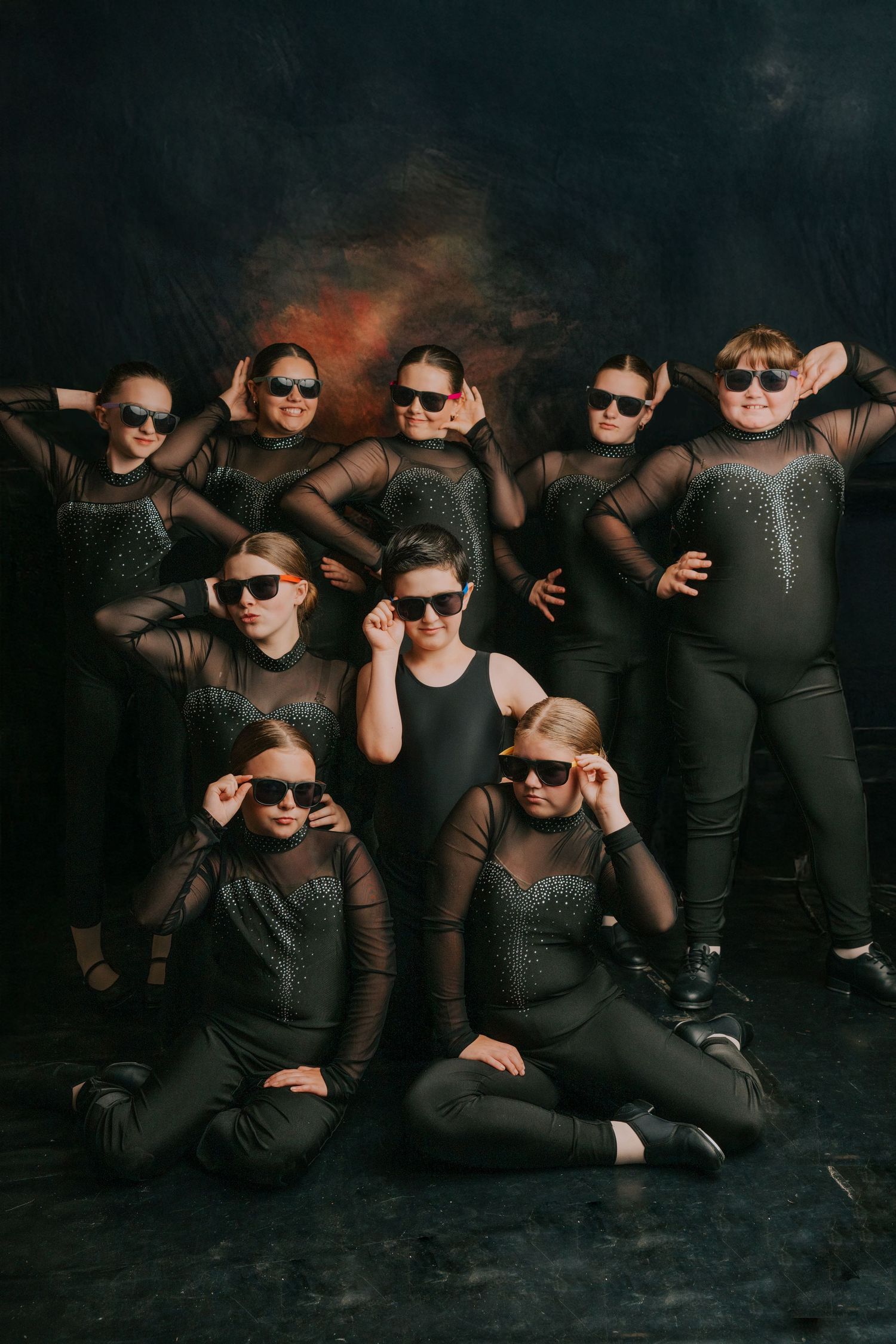 Group of performers in black outfits and sunglasses pose artistically on dark floor in moody lighting.