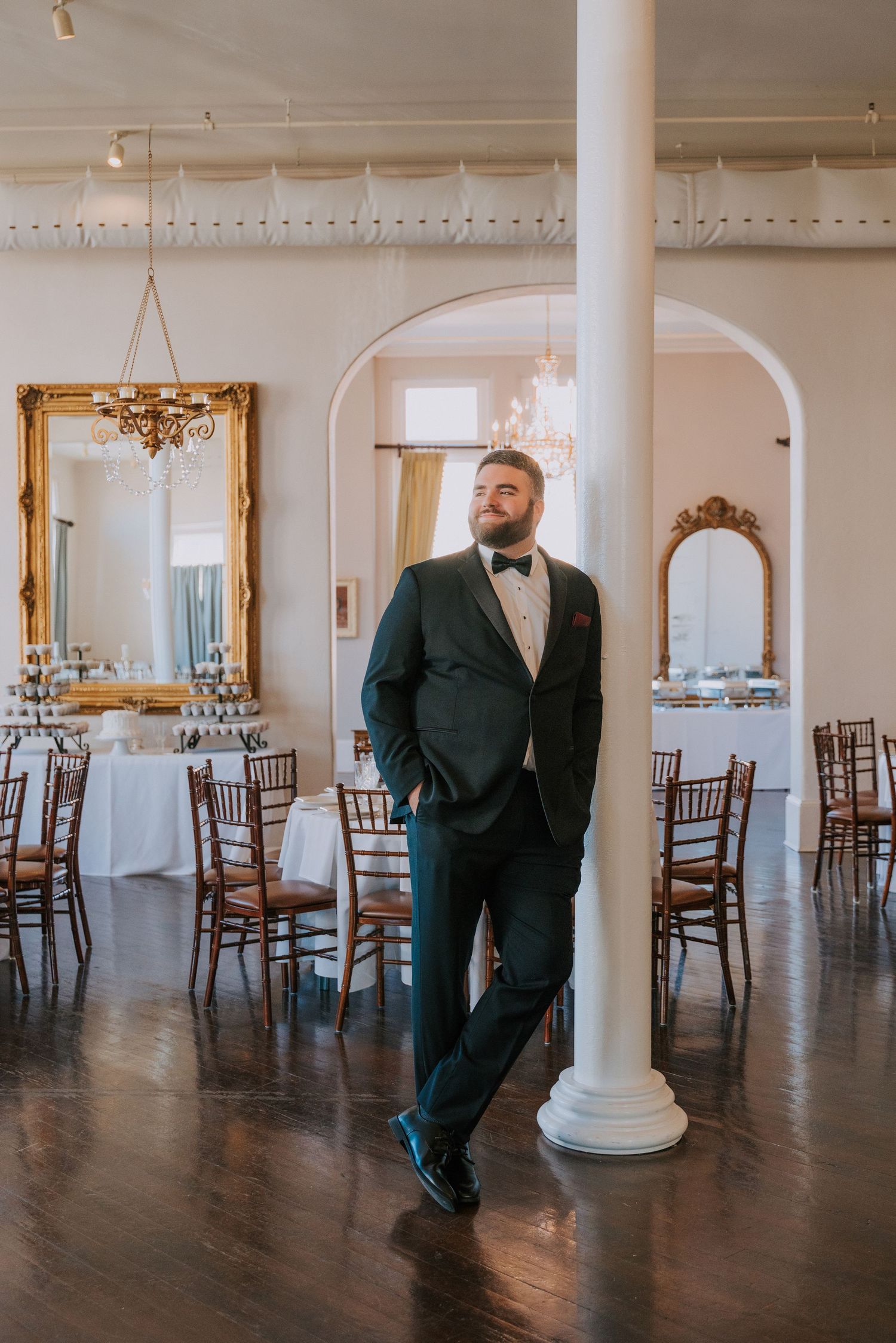 An elegant reception venue with hardwood floors, ornate gold mirrors, white columns and chiavari chairs set for a wedding.
