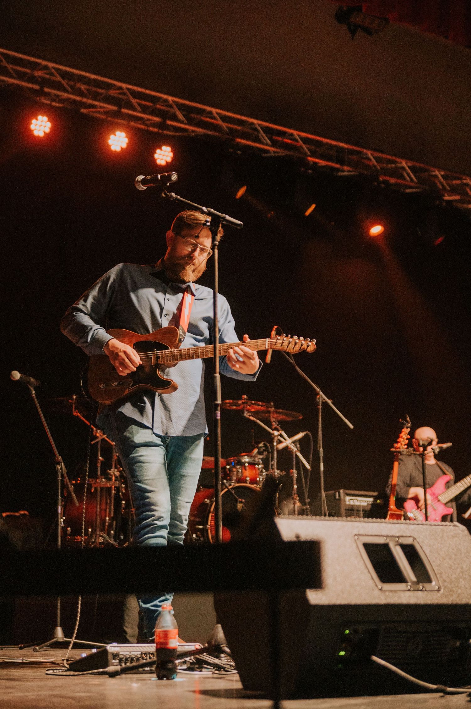A musician performs on stage with a guitar under warm red lighting at a concert venue with stage equipment visible.