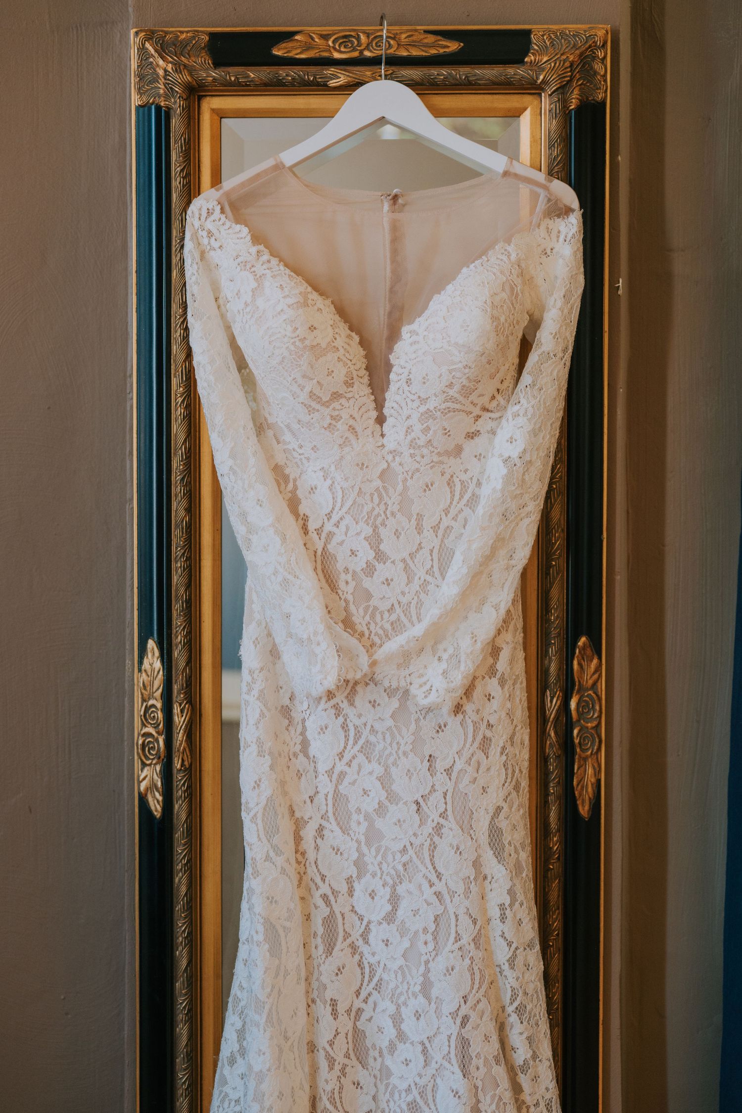 Elegant white lace wedding dress with plunging neckline and long sleeves hanging on display against ornate gold frame mirror.