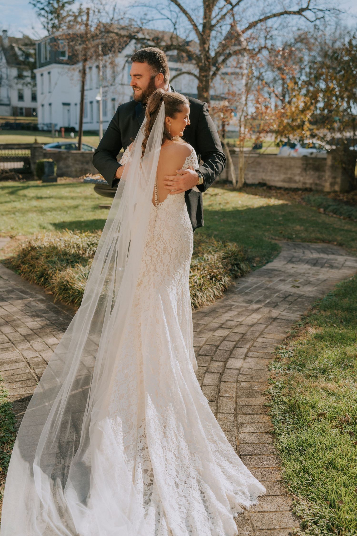 A couple embraces outdoors on a curved stone path while wearing elegant wedding attire with a flowing lace veil trailing behind.