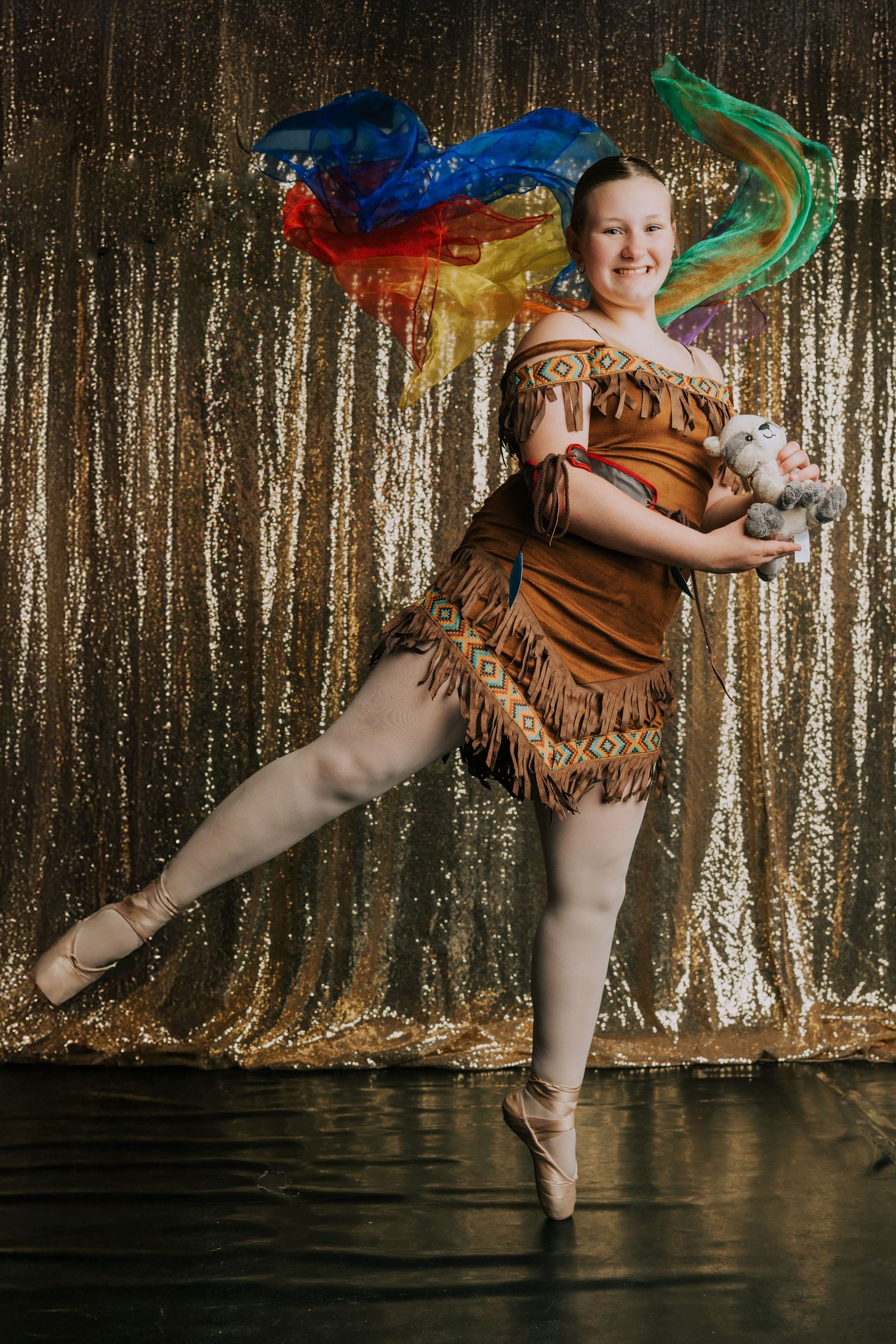 Dancer in brown costume with colorful props performs against gold sequin backdrop.