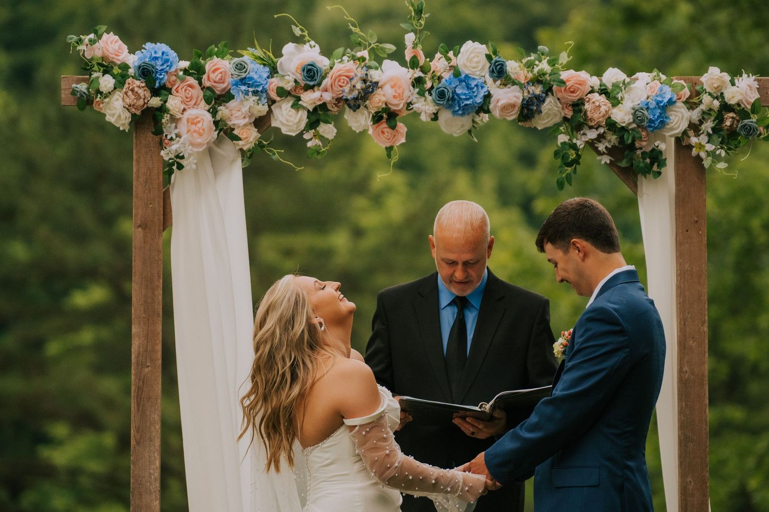 Wooden wedding arch decorated with pink and blue flowers against a mountain landscape.